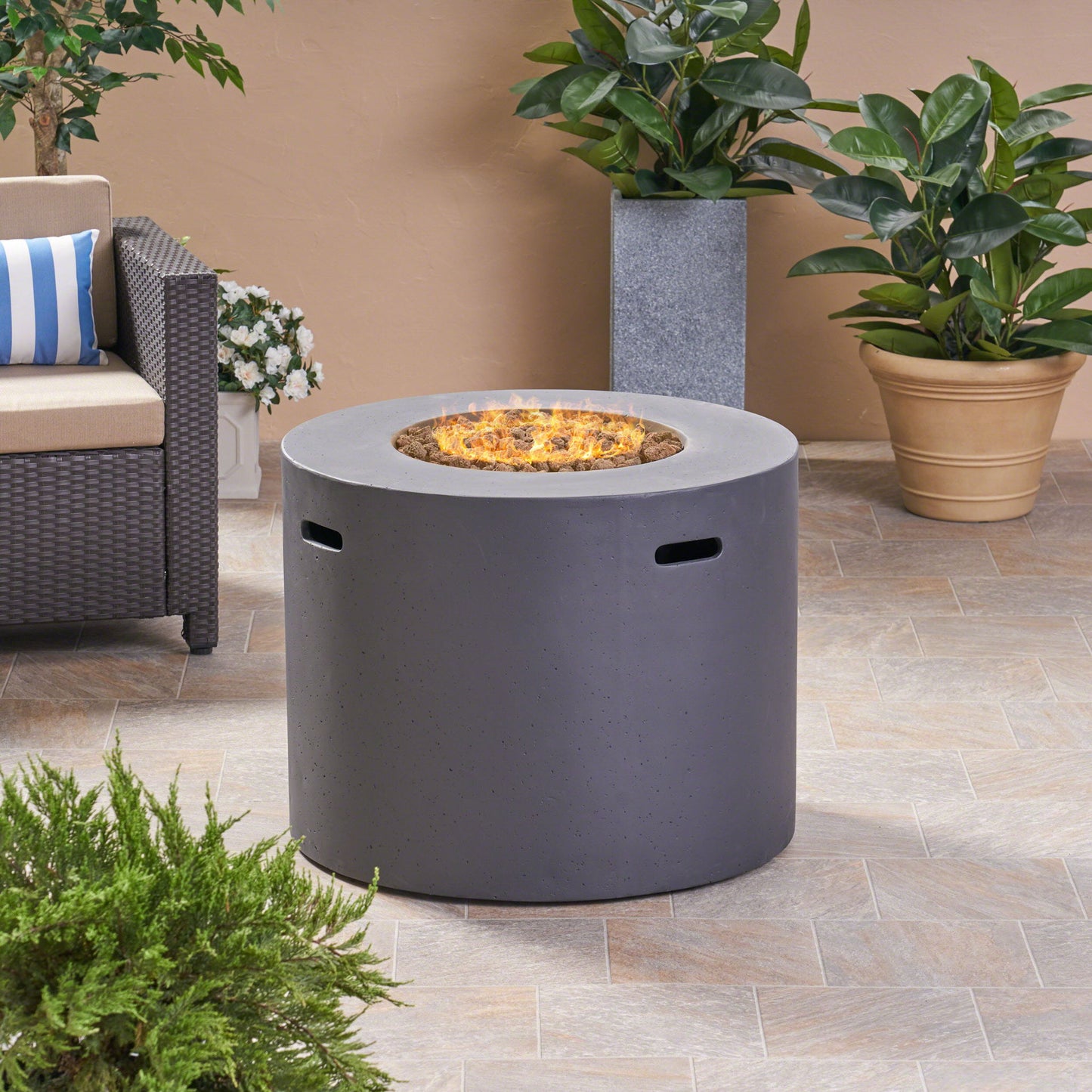 A modern Round Iron Fire Pit With 40,000 Btu sits at the center with flames, surrounded by potted green plants, a sofa with striped cushions, and a tiled patio floor.