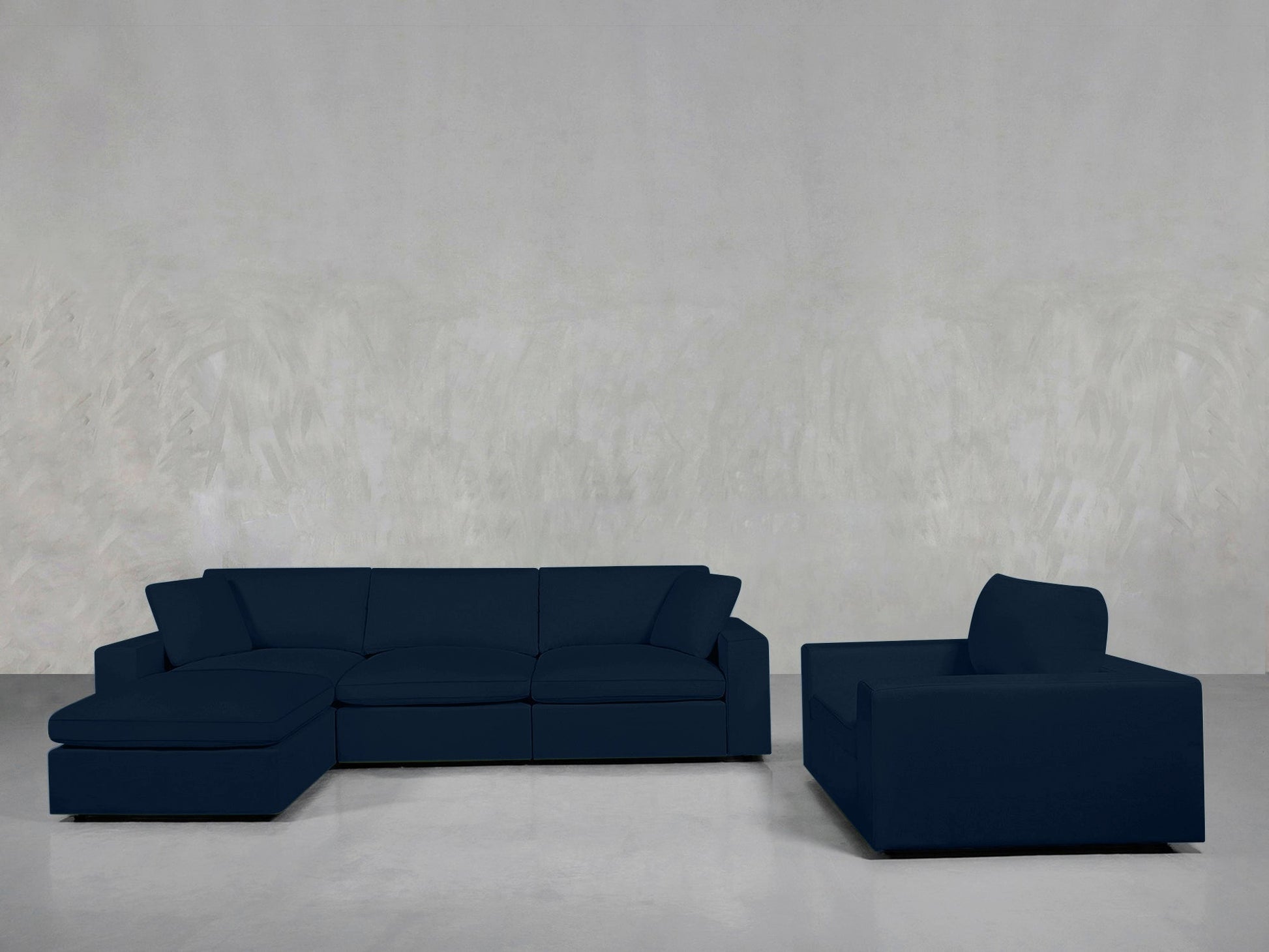 The 4-1 Sectional & Armchair Set, featuring a modern dark blue sectional with chaise and a matching armchair, enhances a minimalist room with light gray walls and flooring.