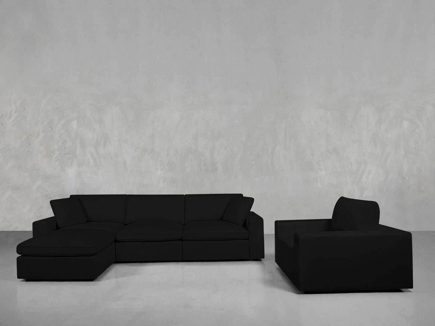 A modern living room features the 4-1 Sectional & Armchair Set—a black sectional with a left-side chaise and matching armchair—arranged against a light gray textured wall and gray flooring.