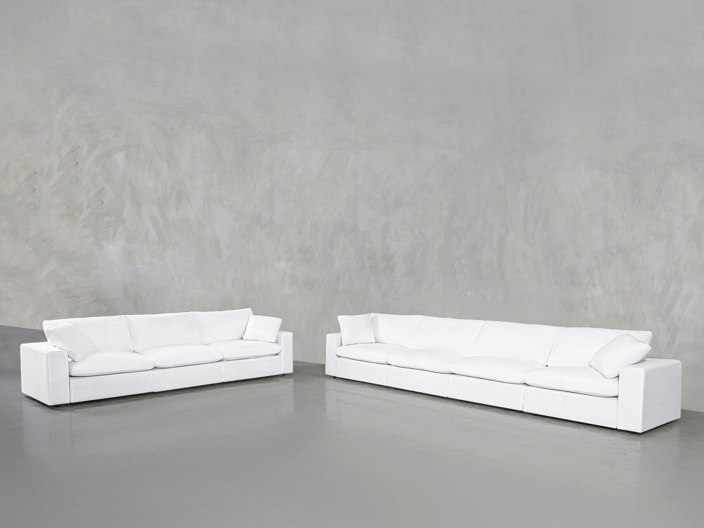 The 4-3 Sofa Set, featuring modern white sofas with plush cushions, is arranged on a smooth gray floor against a textured light gray wall in a spacious, minimalist setting.
