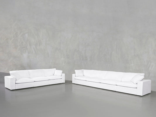 The 4-3 Sofa Set, featuring modern white sofas with plush cushions, is arranged on a smooth gray floor against a textured light gray wall in a spacious, minimalist setting.