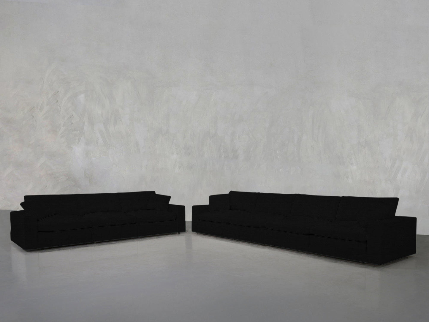 The minimalistic room features the 4-3 Sofa Set—two large, black sofas arranged in an L-shape against a plain, light gray textured wall on a smooth, light-colored floor. No other furniture is present.