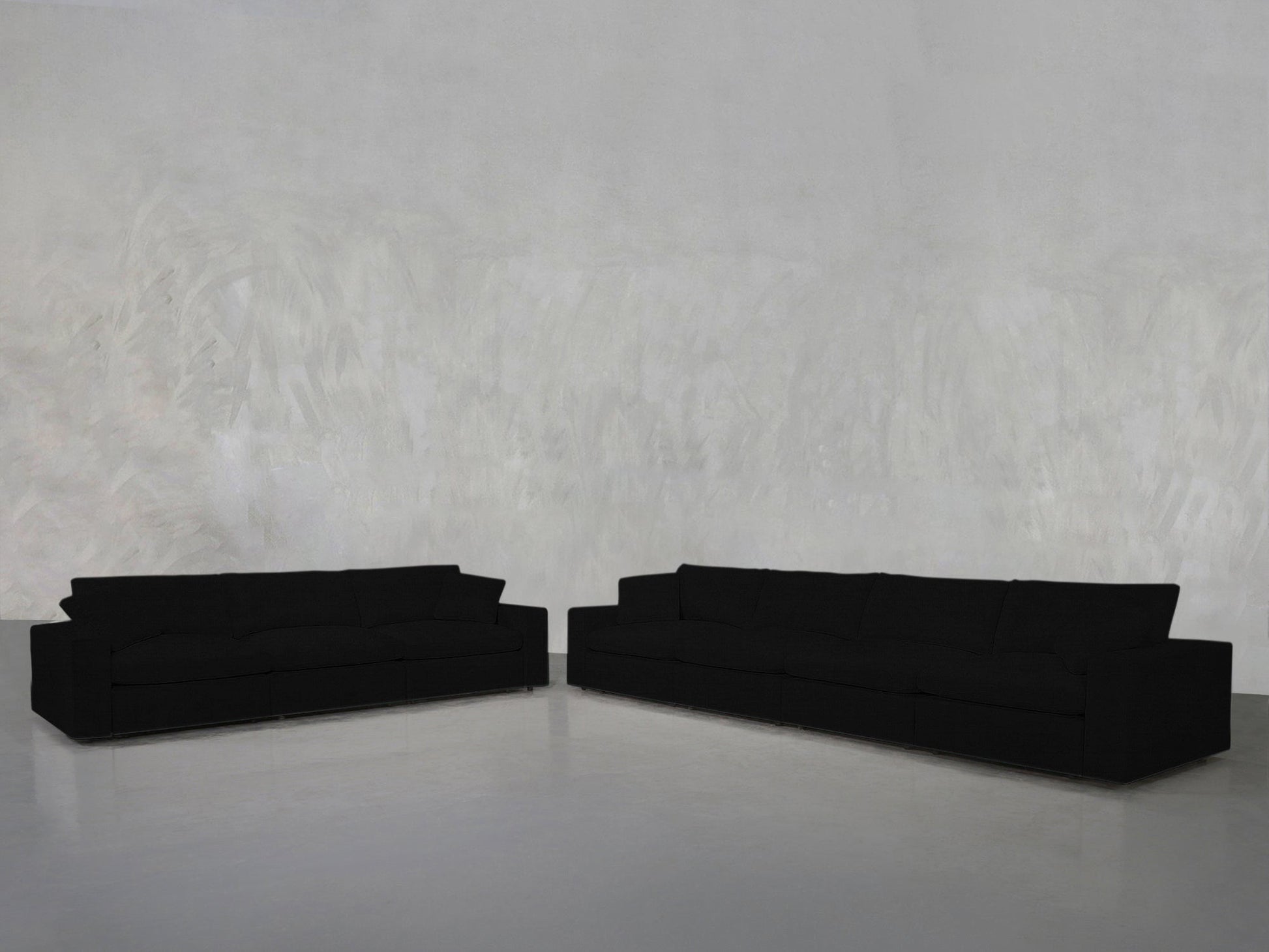The minimalistic room features the 4-3 Sofa Set—two large, black sofas arranged in an L-shape against a plain, light gray textured wall on a smooth, light-colored floor. No other furniture is present.