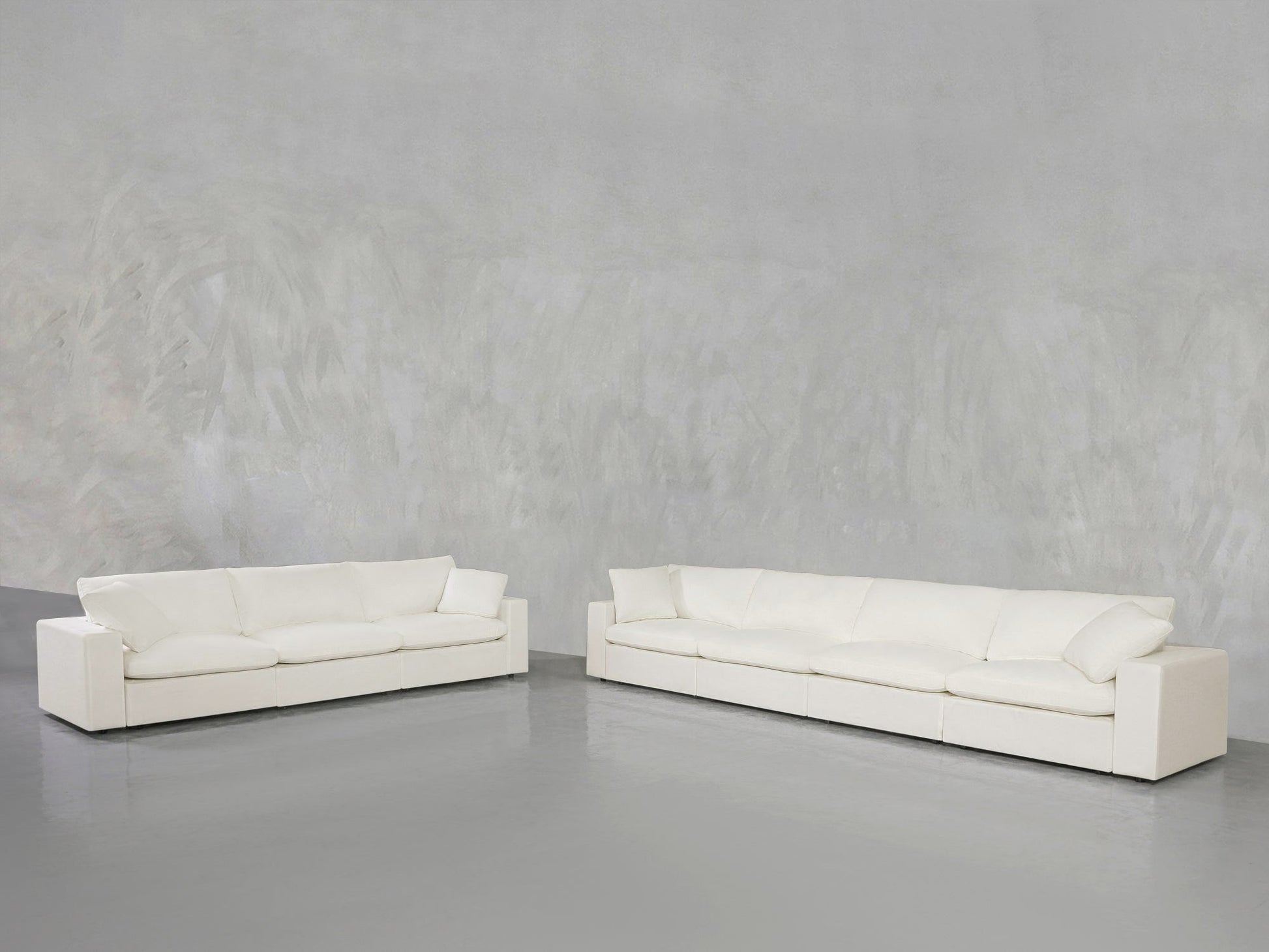 The 4-3 Sofa Set in modern white with large cushions is arranged in an L-shape against a plain, light gray textured wall on a smooth, gray floor.