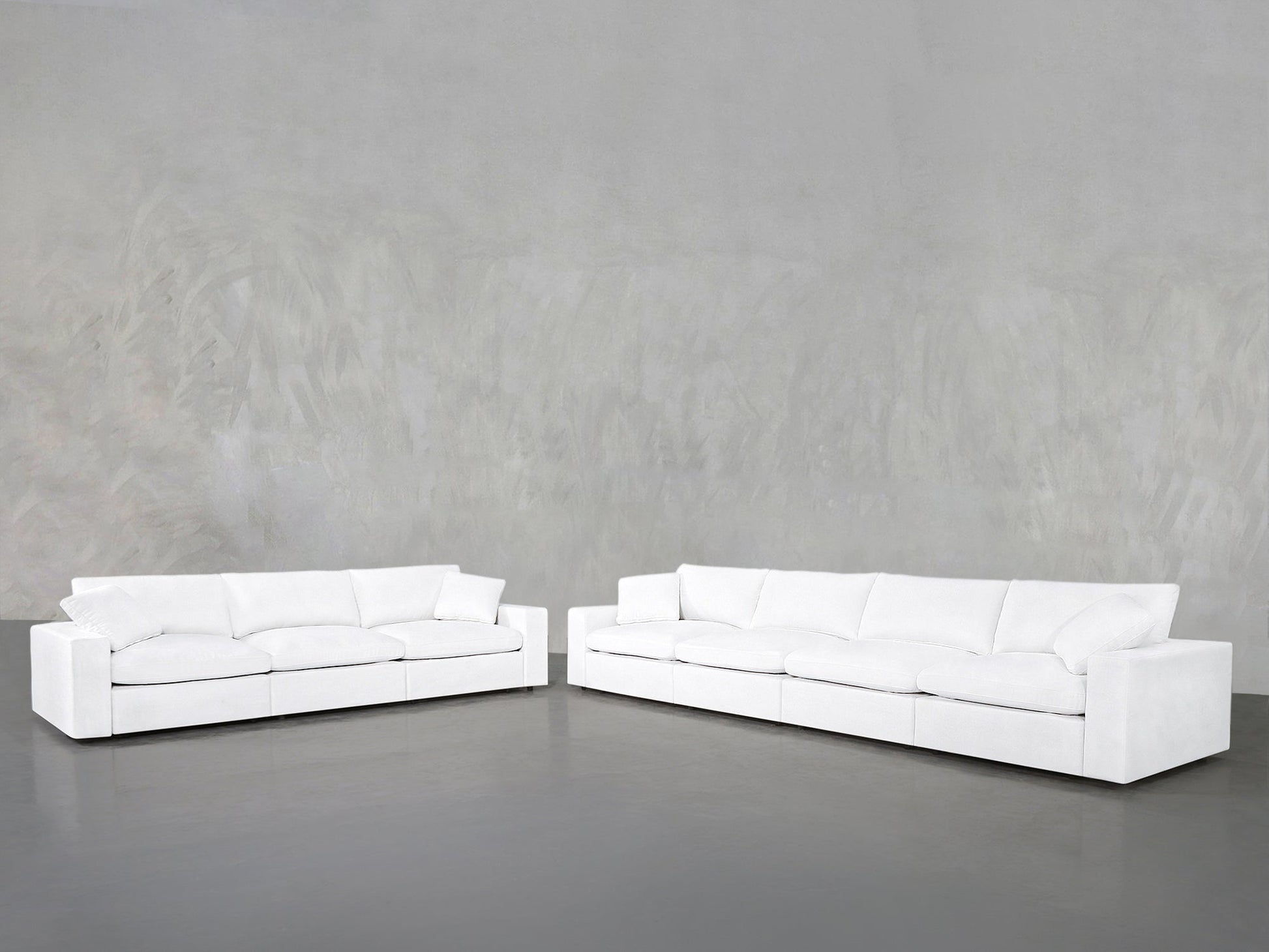 The 4-3 Sofa Set, featuring two modern white sofas with plush cushions, is arranged in an L-shape against a light gray wall and dark gray floor for a minimalistic, spacious look.