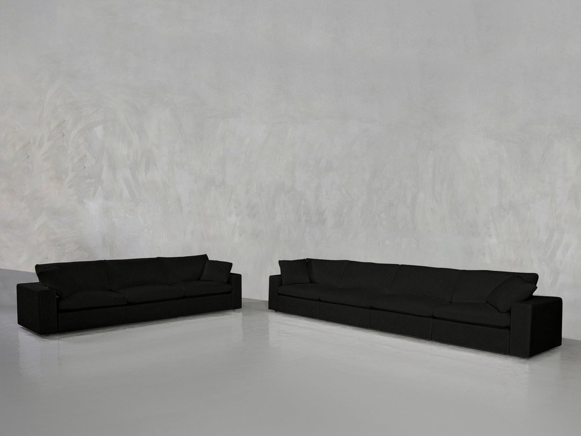 The 4-3 Sofa Set, featuring two long black sofas with clean, modern lines, sits on a shiny light gray floor against a textured pale gray wall in a minimalist, spacious room.
