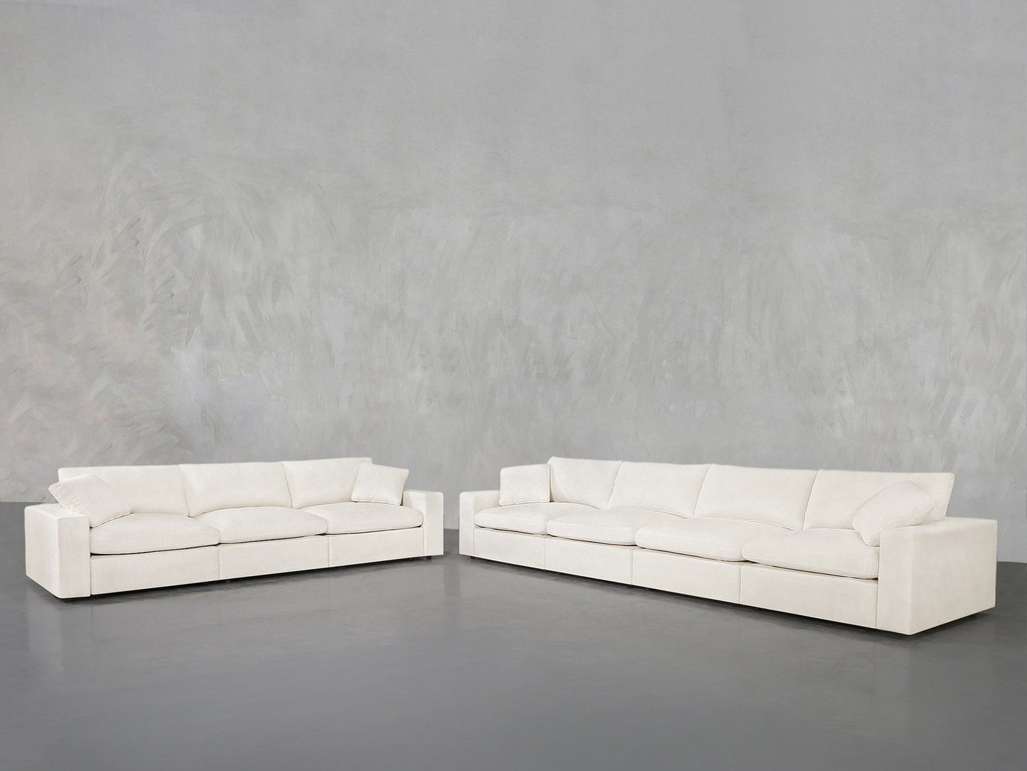 The 4-3 Sofa Set, featuring two modern white sofas with clean lines and plush cushions, is arranged in an L-shape against a plain light gray wall and floor for a minimalist, spacious look.