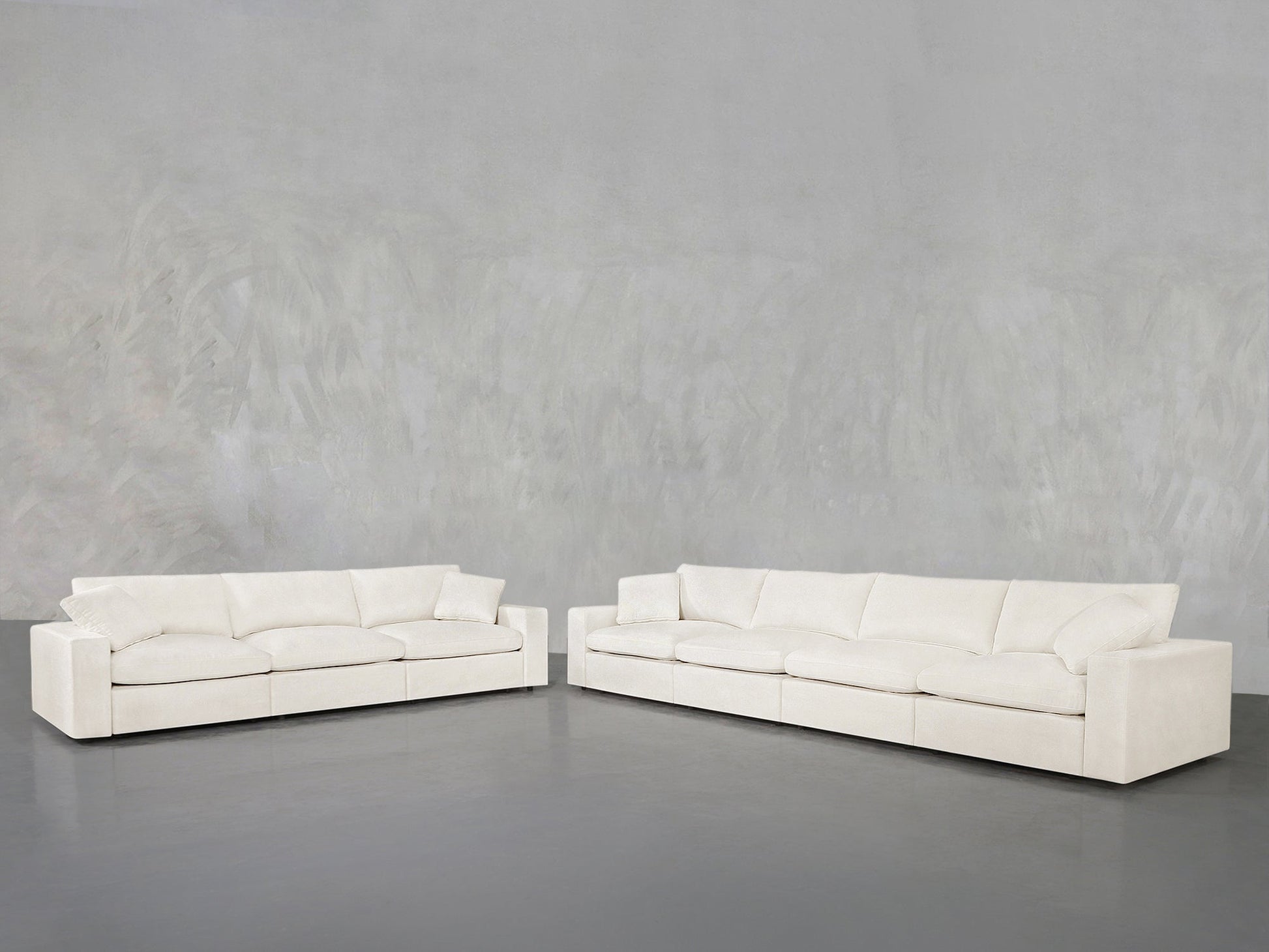 The 4-3 Sofa Set, featuring two modern white sofas with clean lines and plush cushions, is arranged in an L-shape against a plain light gray wall and floor for a minimalist, spacious look.