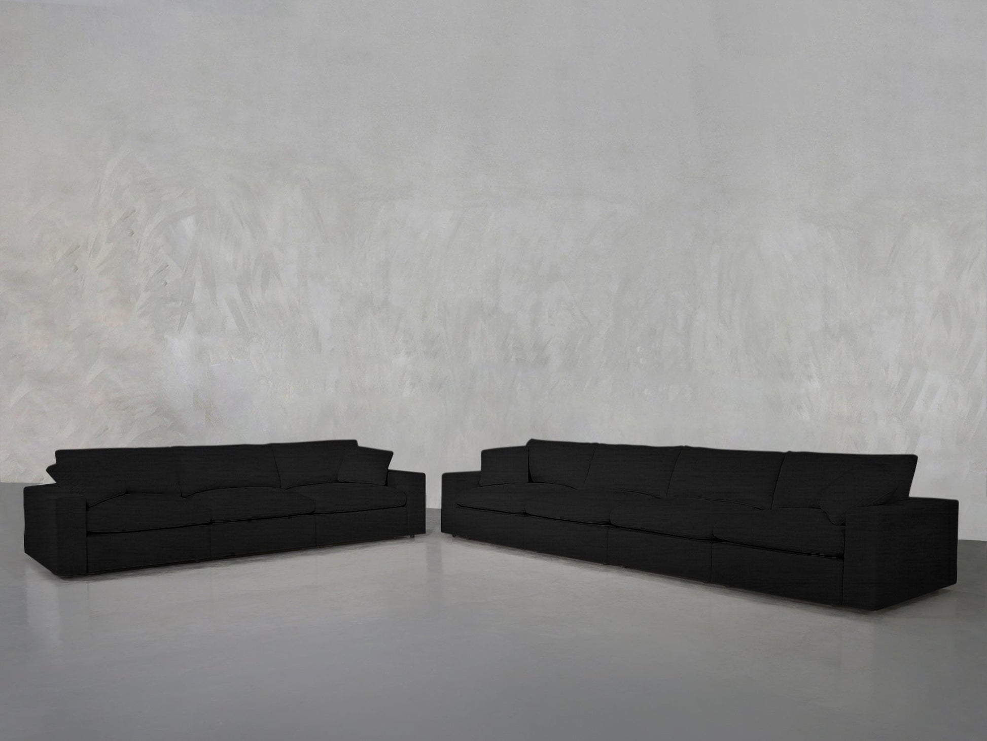 The 4-3 Sofa Set, featuring a modern black design, is arranged at an angle in an empty room with a light gray textured wall and smooth gray floor to create an inviting corner seating area.