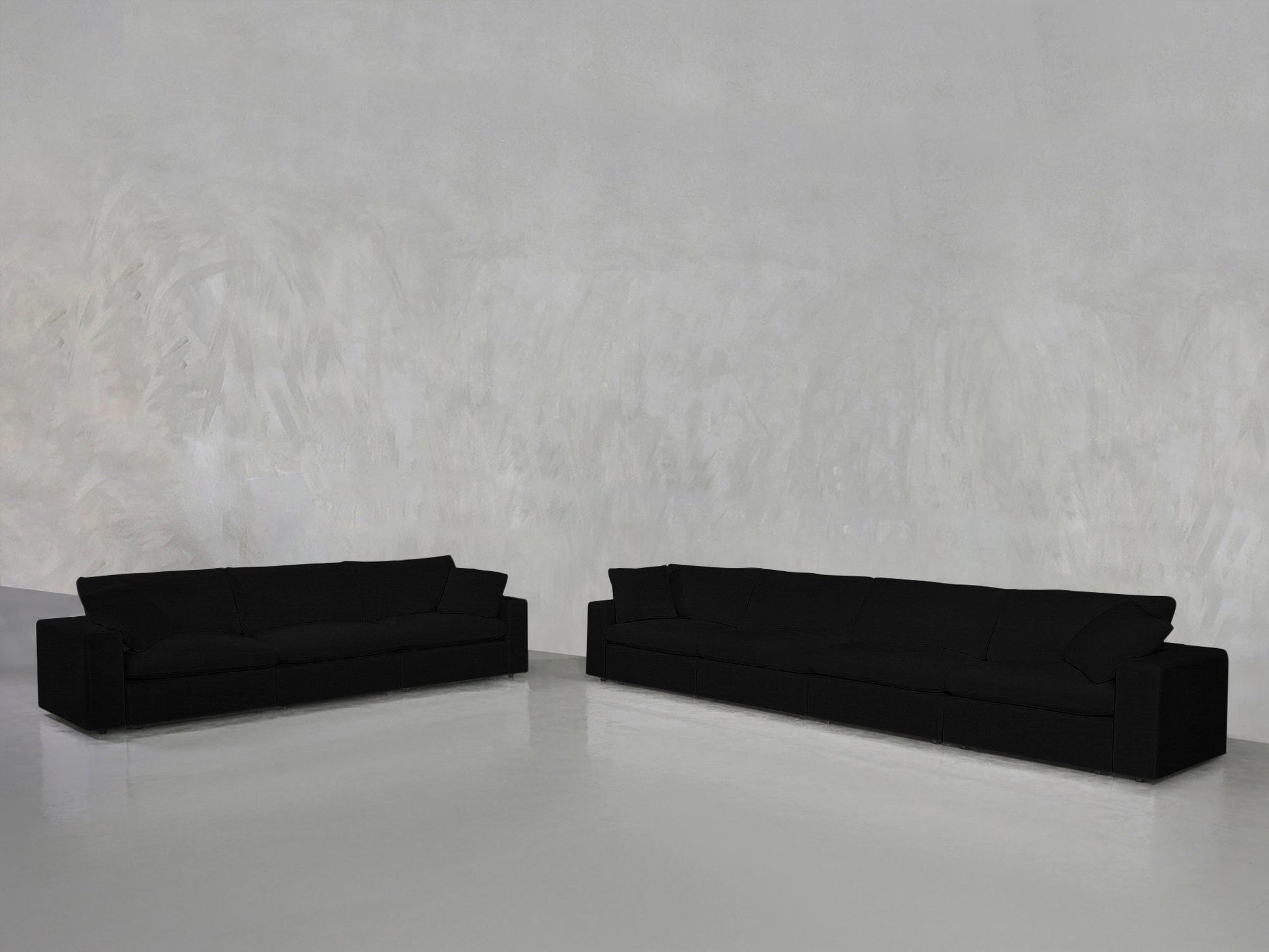 The 4-3 Sofa Set, featuring two black sofas of varying lengths, is arranged on a polished light gray floor against a textured light gray wall in a minimalist setting.