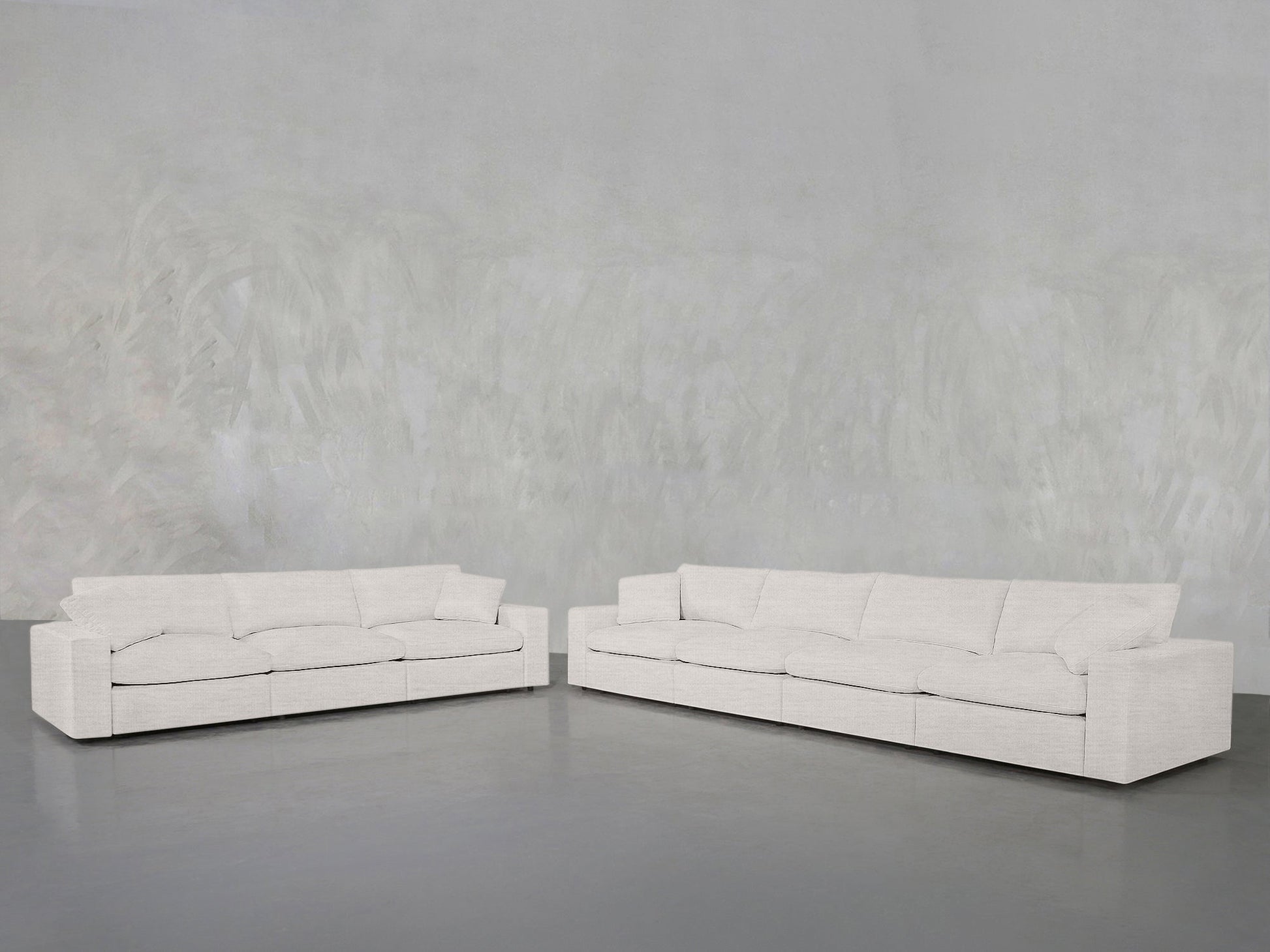 The 4-3 Sofa Set, a modern light-colored sectional with plush cushions, is arranged in an L-shape in a spacious minimalist room featuring a textured gray wall and dark gray floor.