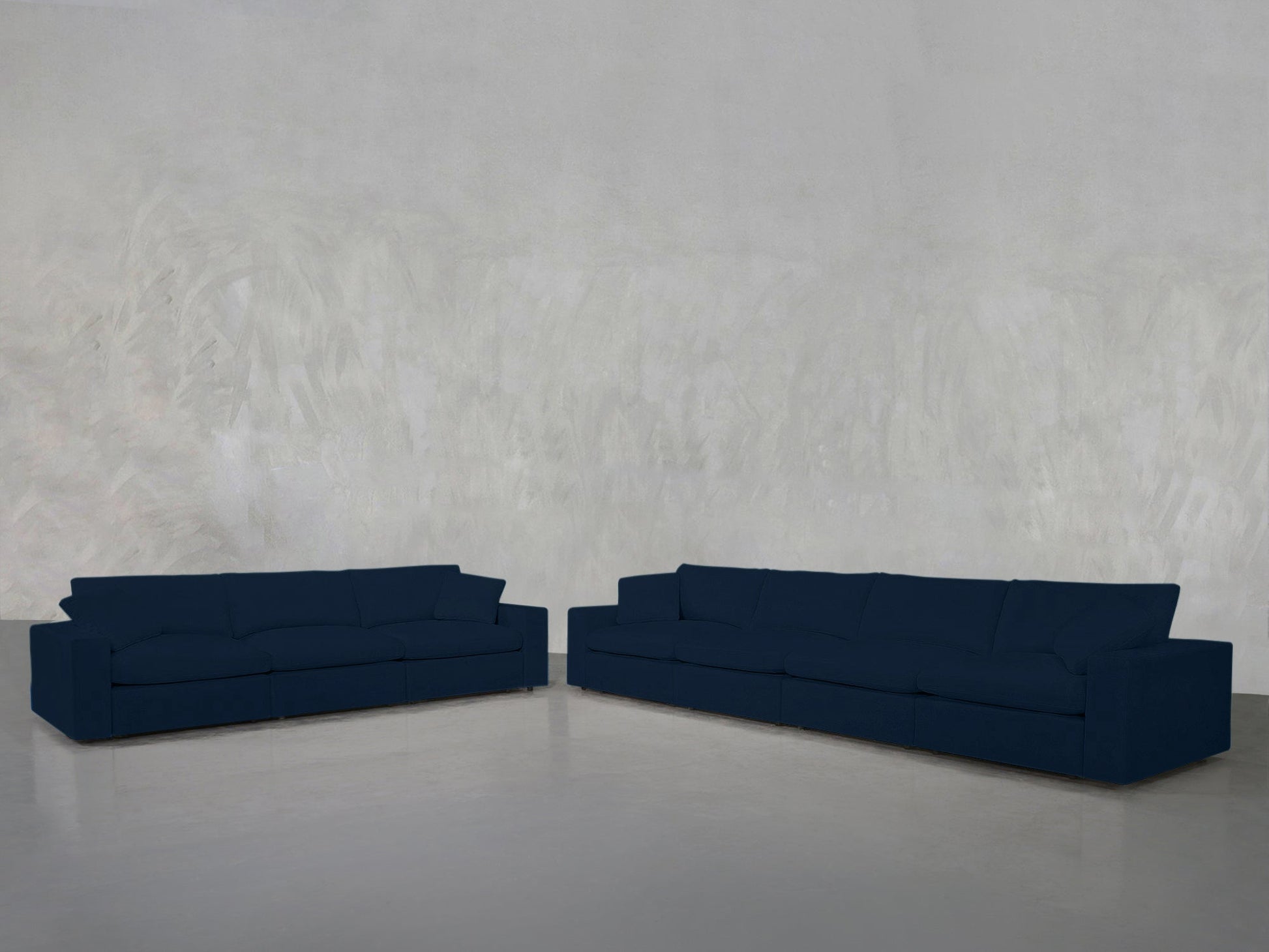 The 4-3 Sofa Set features two modern dark blue sofas arranged in an L-shape, placed against a plain light gray textured wall and a smooth gray floor.