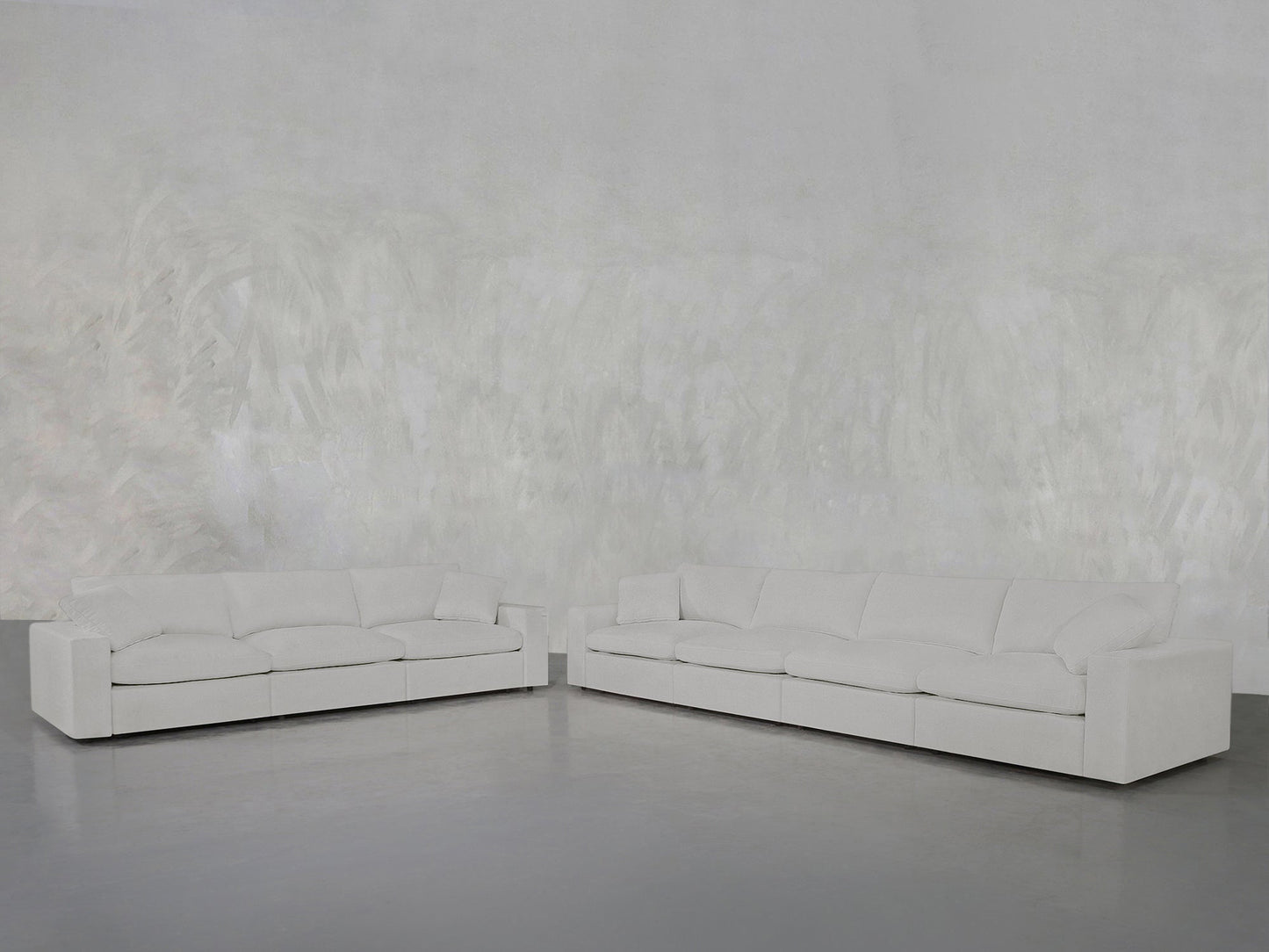 The 4-3 Sofa Set, featuring modern white sofas with plush cushions, sits on a smooth gray floor against a textured light gray wall in a minimalist, empty room.