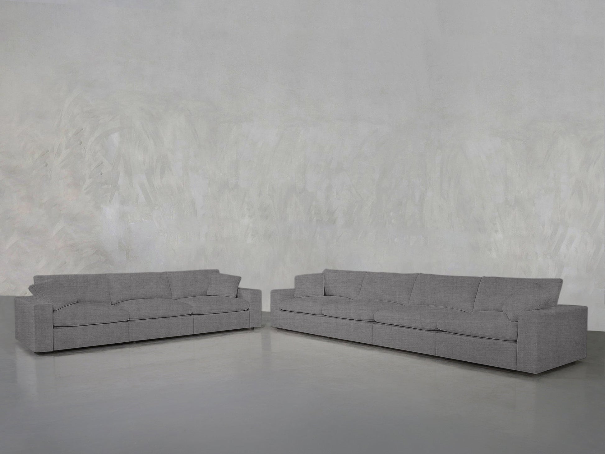 The 4-3 Sofa Set, featuring modern gray upholstery and straight lines, is arranged in an L-shape against a light gray textured wall on a smooth, light-colored floor.