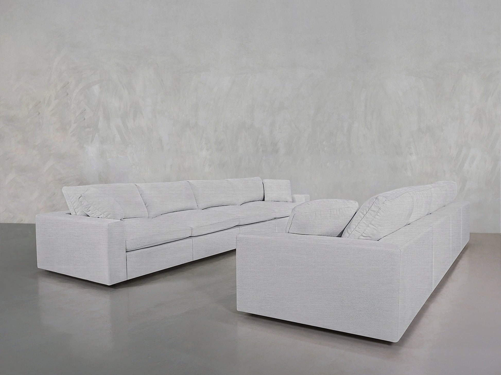 The 4-4 Sofa Set features two light gray, modern sectionals arranged in an L-shape on a polished gray floor, set against a textured pale gray wall. Clean lines and plush cushions add to its contemporary appeal.