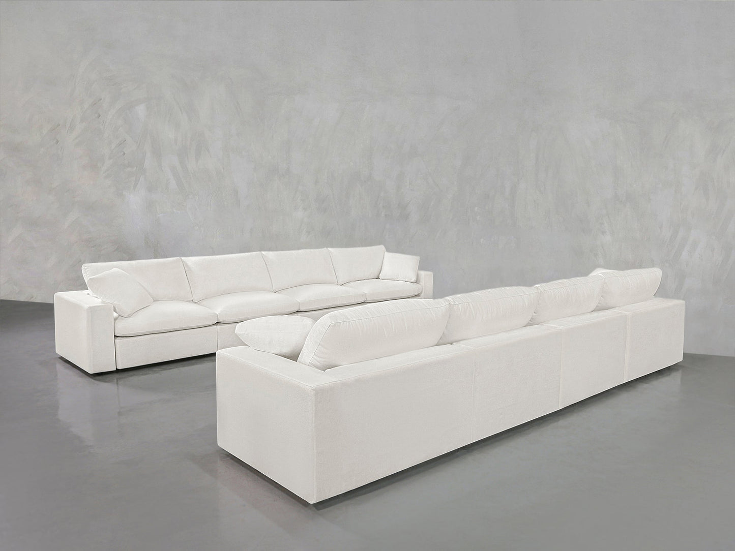 Two 4-4 Sofa Sets with plush cushions are placed facing each other in a minimalist room featuring light gray textured walls and a glossy gray floor.