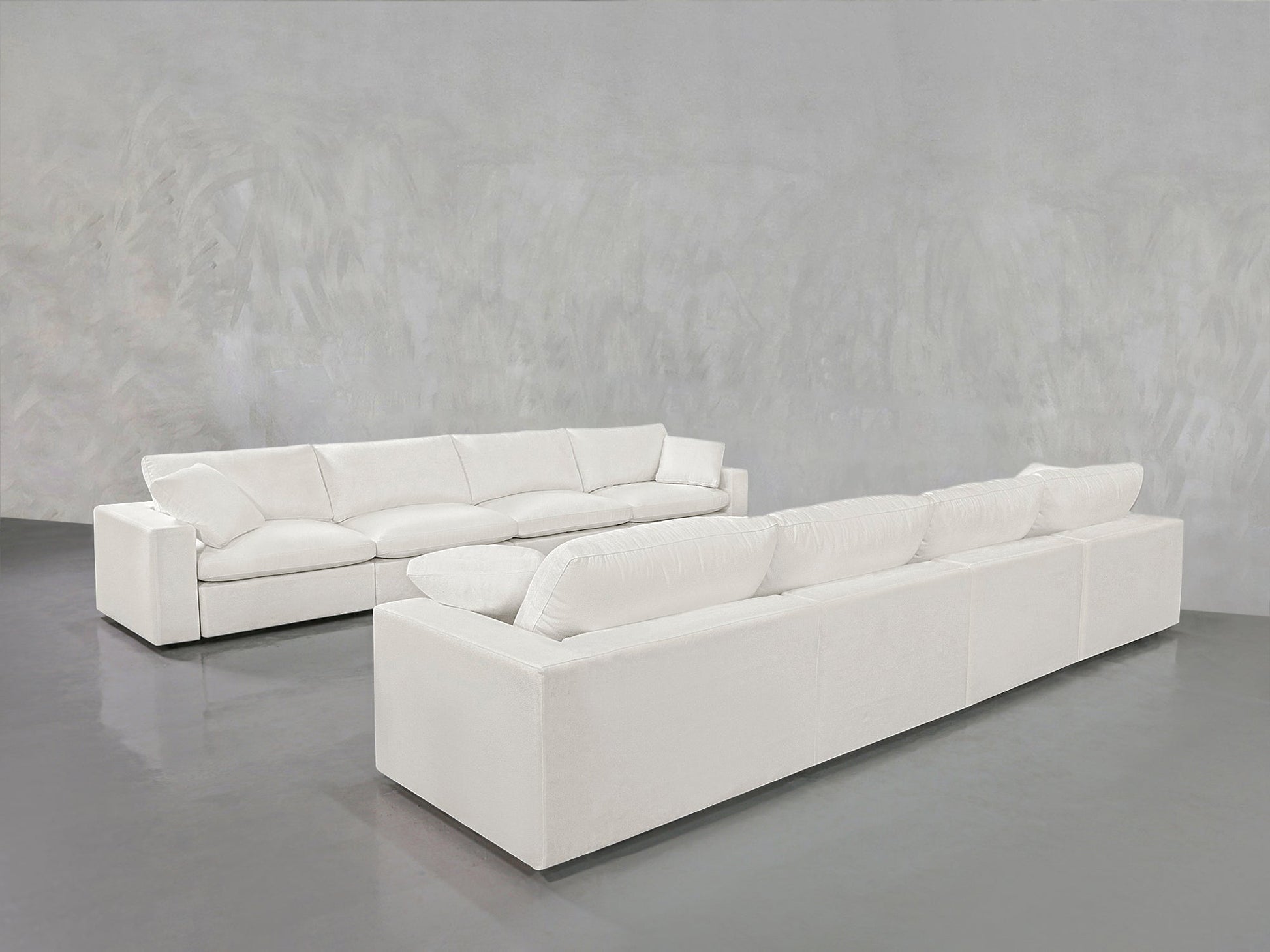 Two 4-4 Sofa Sets with plush cushions are placed facing each other in a minimalist room featuring light gray textured walls and a glossy gray floor.