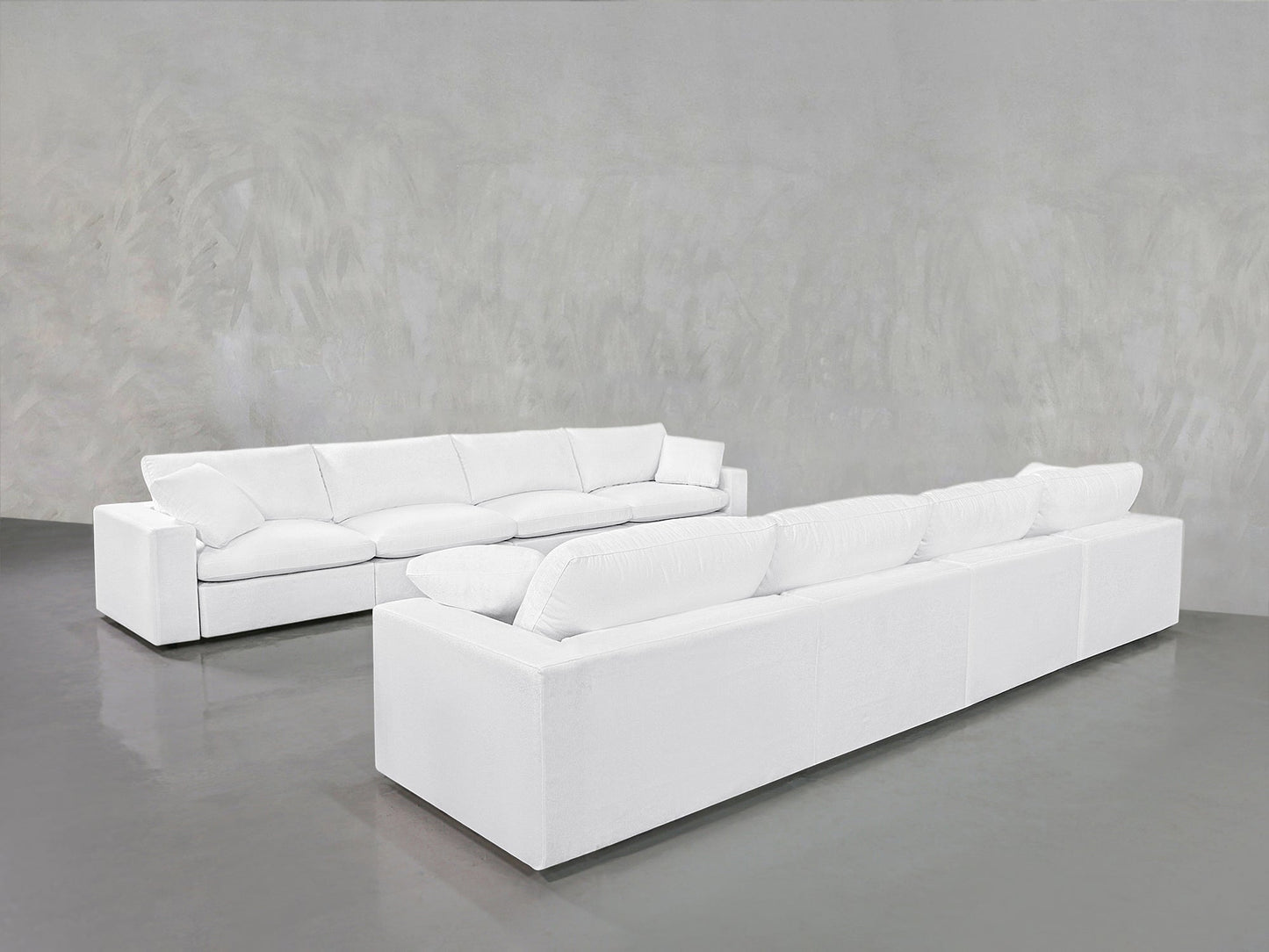 The 4-4 Sofa Set, featuring modern white sofas with plush cushions, is arranged in an L-shape on a smooth gray floor against a textured light gray wall in a spacious, minimalist room.