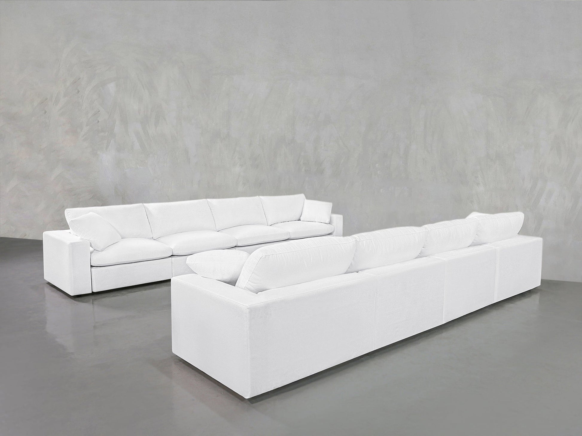 The 4-4 Sofa Set, featuring modern white sofas with plush cushions, is arranged in an L-shape on a smooth gray floor against a textured light gray wall in a spacious, minimalist room.