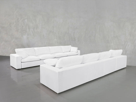 The 4-4 Sofa Set, featuring modern white sofas with plush cushions, is arranged in an L-shape on a smooth gray floor against a textured light gray wall in a spacious, minimalist room.