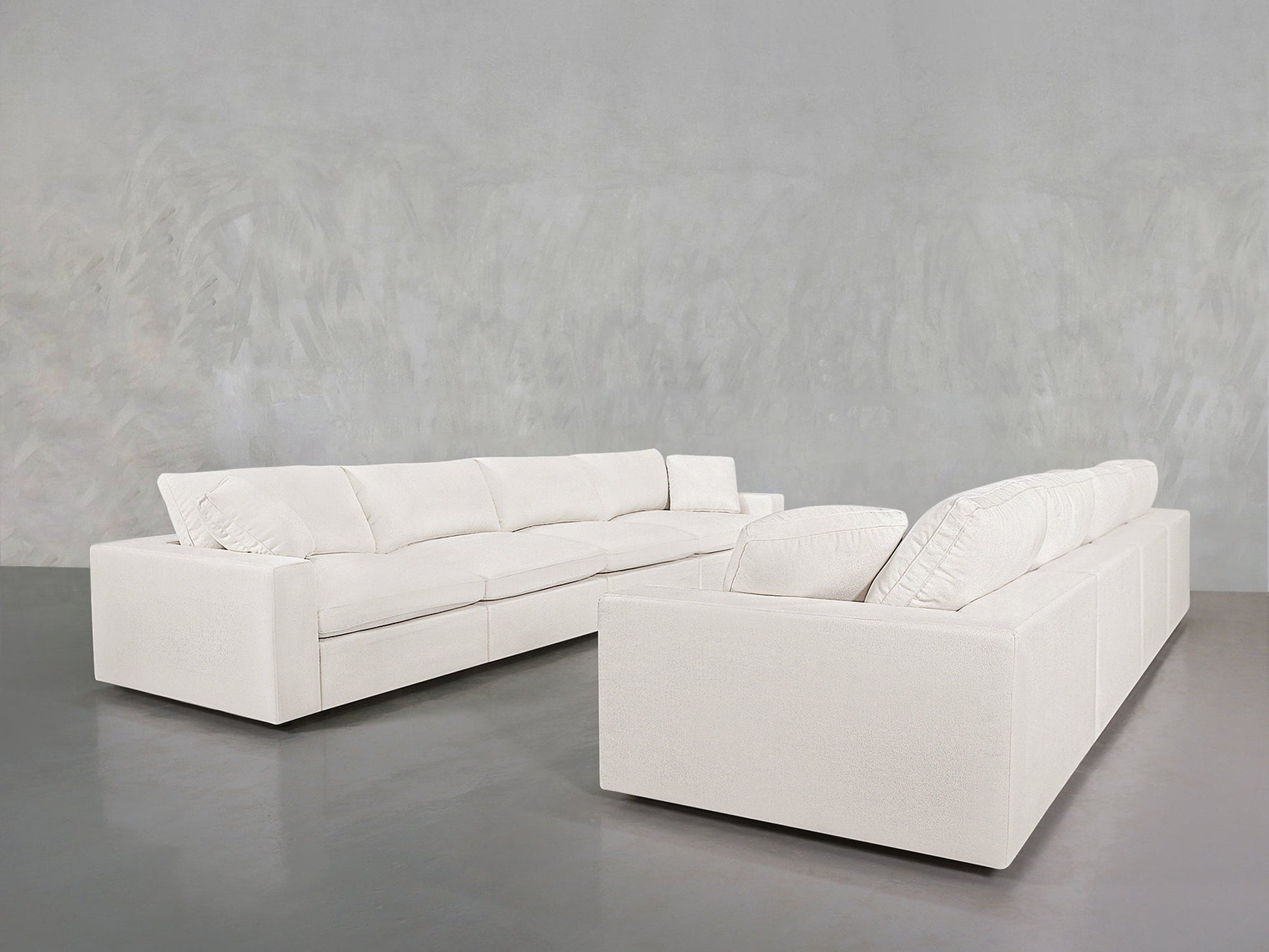 Two 4-4 Sofa Sets with clean lines and plush cushions are arranged on a gray polished floor against a light gray textured wall.