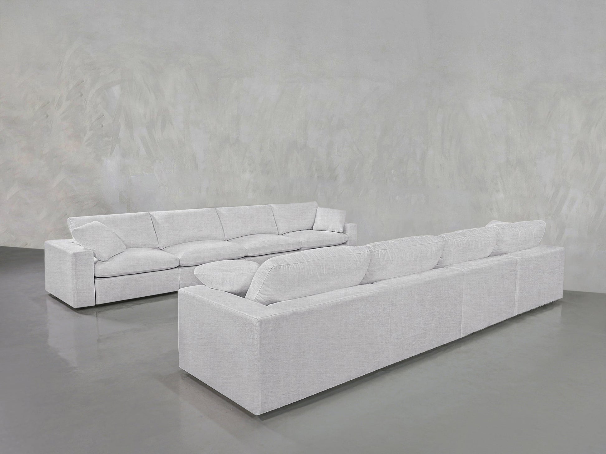 Two 4-4 Sofa Sets with plush cushions are arranged facing each other in a minimalistic, light gray room featuring a polished concrete floor and textured walls.