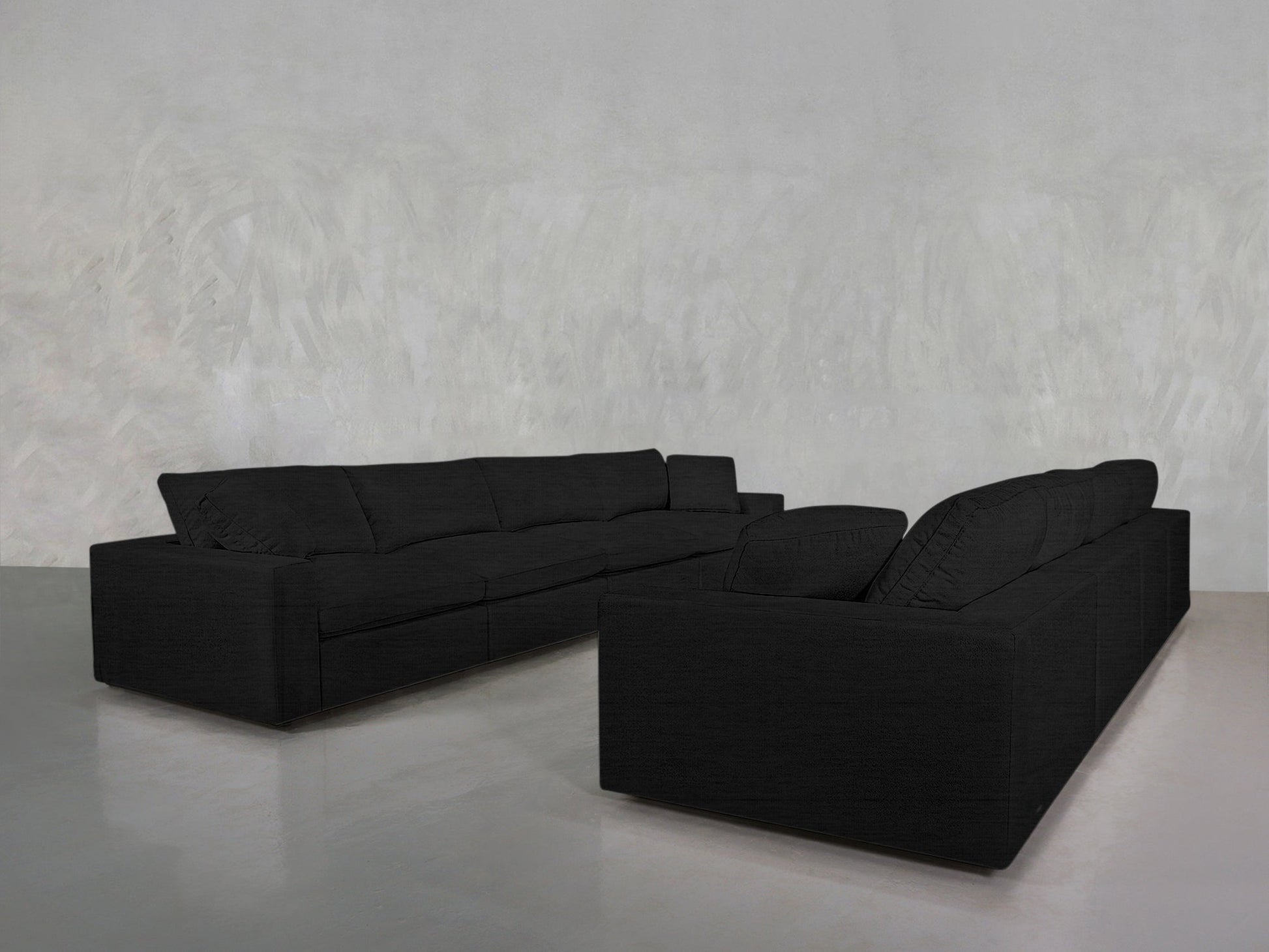 The 4-4 Sofa Set, in modern black, is arranged in an L-shape in a minimalistic room with light gray walls and a reflective gray floor.