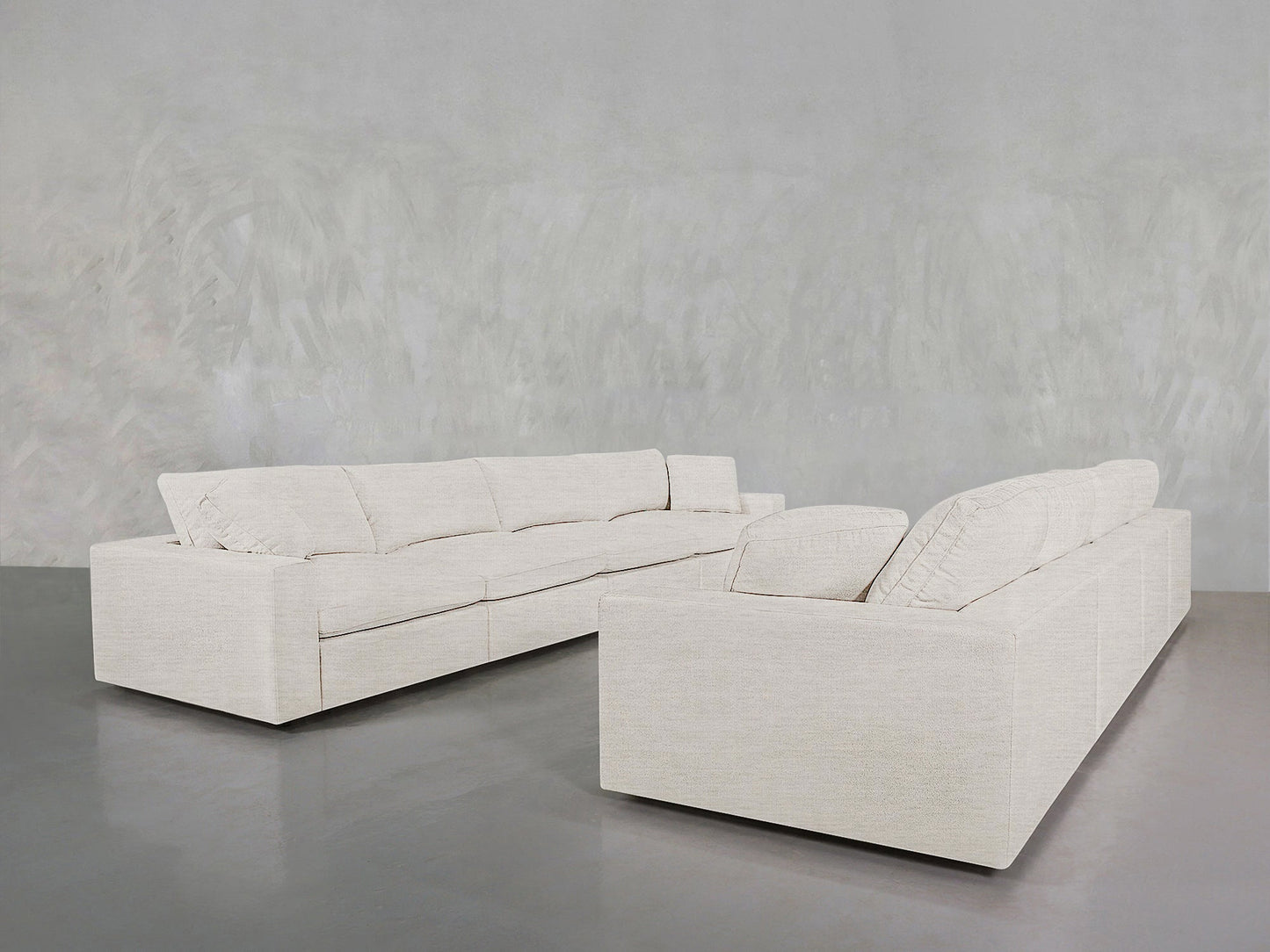 Two large, modern 4-4 Sofa Sets in light beige with plush cushions are arranged L-shaped on a smooth grey floor against a textured grey wall.