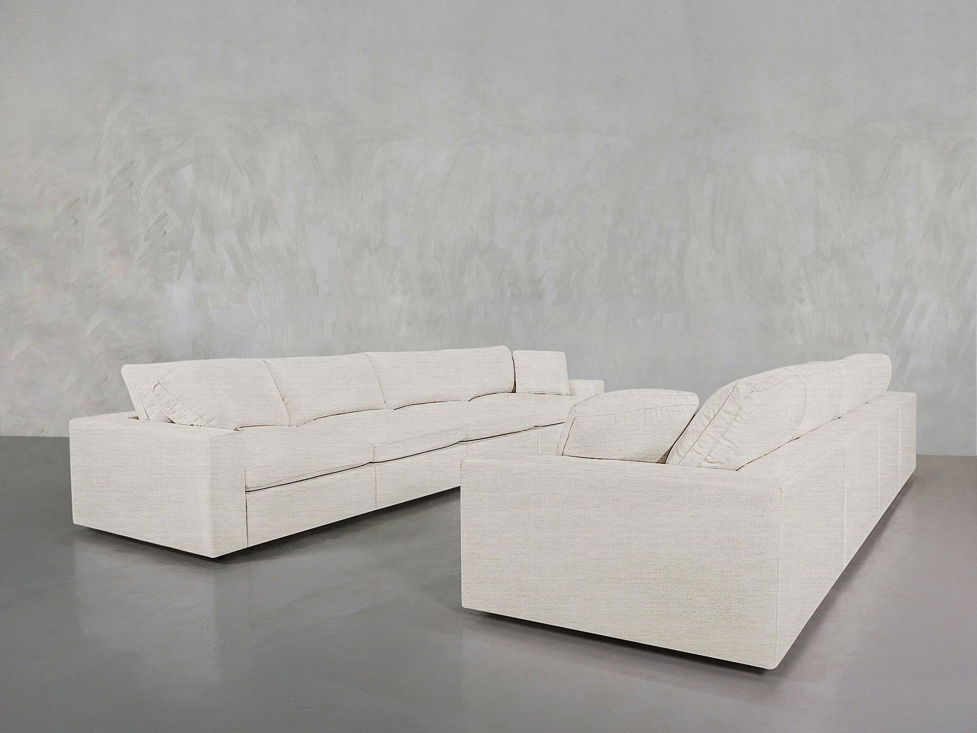 Two large, modern 4-4 Sofa Sets in light beige with plush cushions are arranged L-shaped on a smooth grey floor against a textured grey wall.