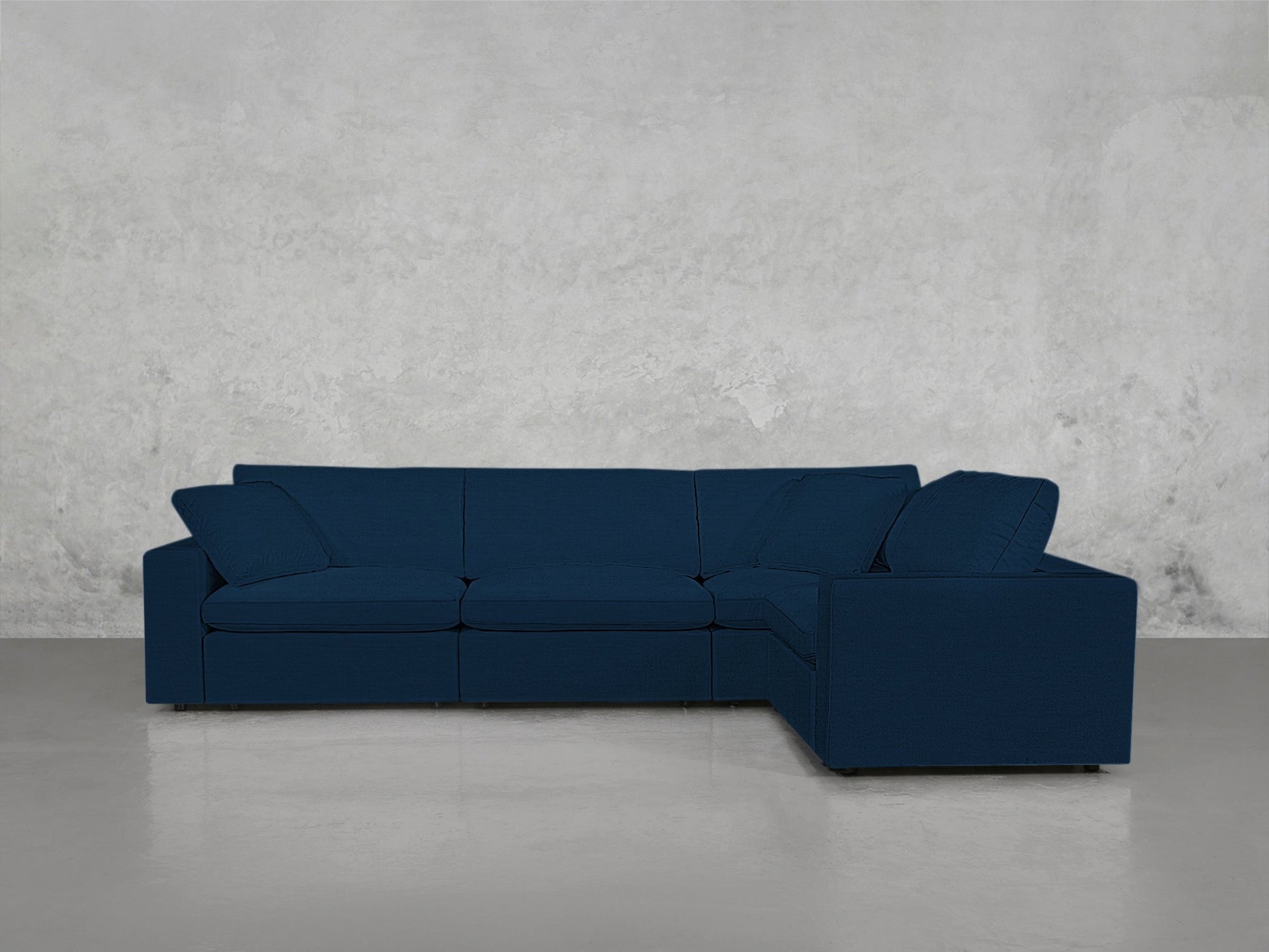 A 4-Seat Modular Corner Sectional in dark blue with clean lines and low backrests is set against a light gray textured wall on a smooth, light-colored floor.