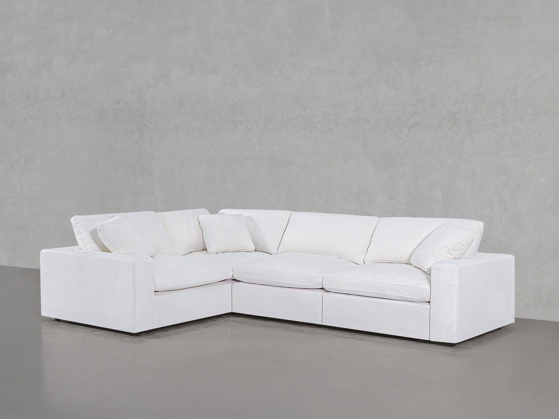 A 4-Seat Modular Corner Sectional with plush cushions and pillows sits on a polished concrete floor against a light gray textured wall.
