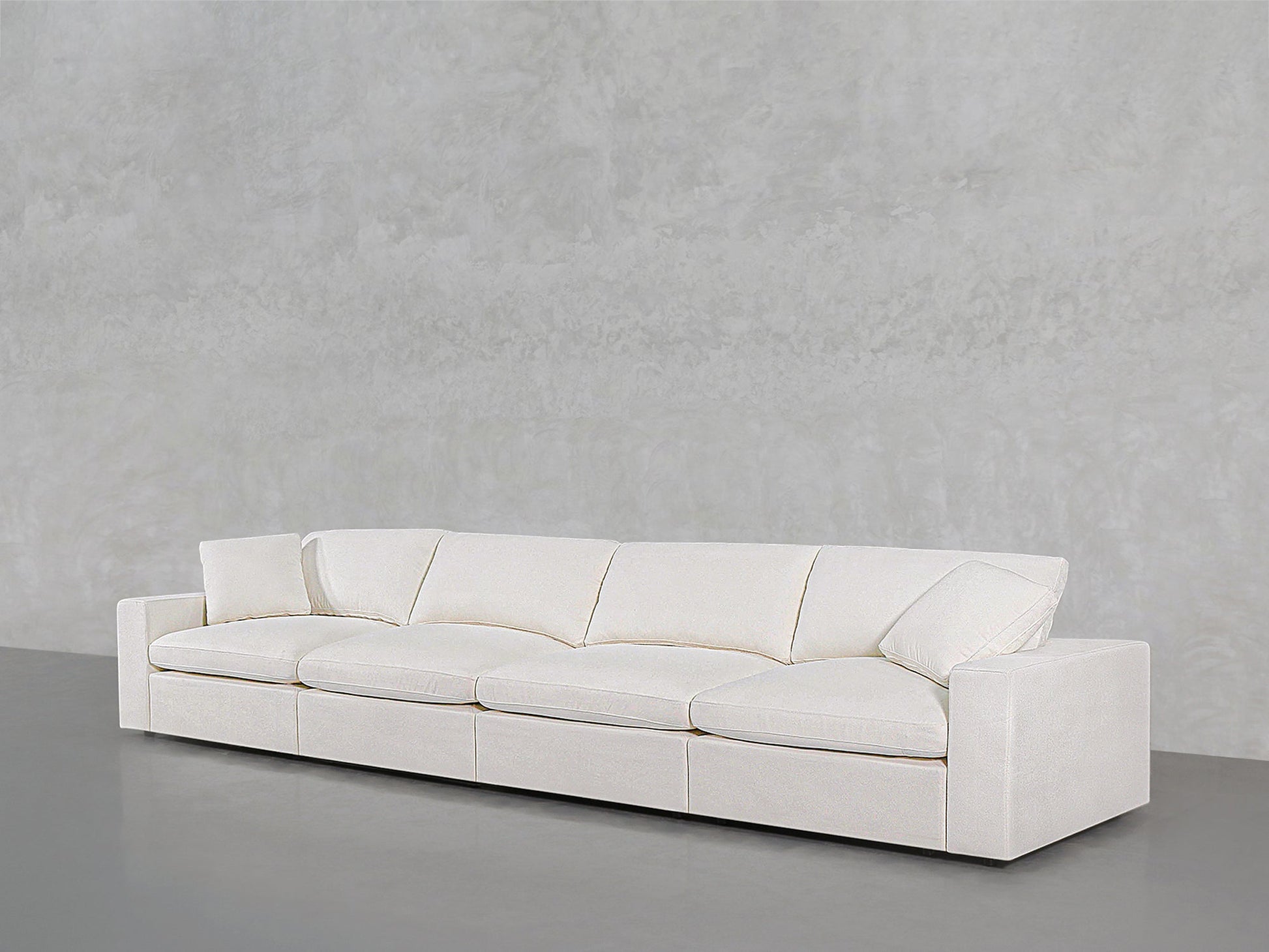 The 4-Seat Modular Sofa, featuring a modern white design with four seat cushions and two angled armrests, is set against a light gray textured wall on a gray floor.