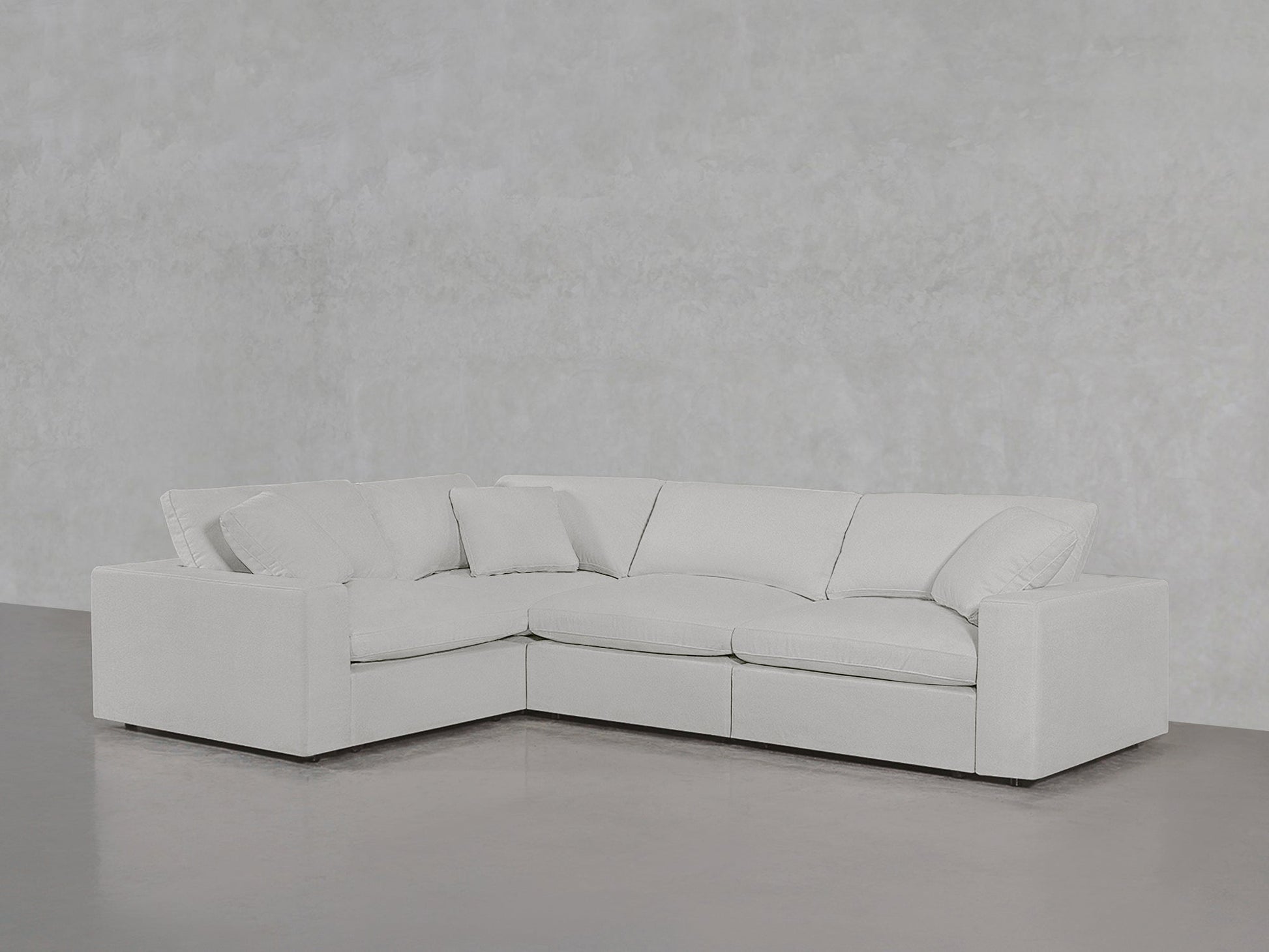 A 4-Seat Modular Corner Sectional in modern light gray with plush cushions sits in a minimalist room featuring light gray walls and a polished concrete floor.