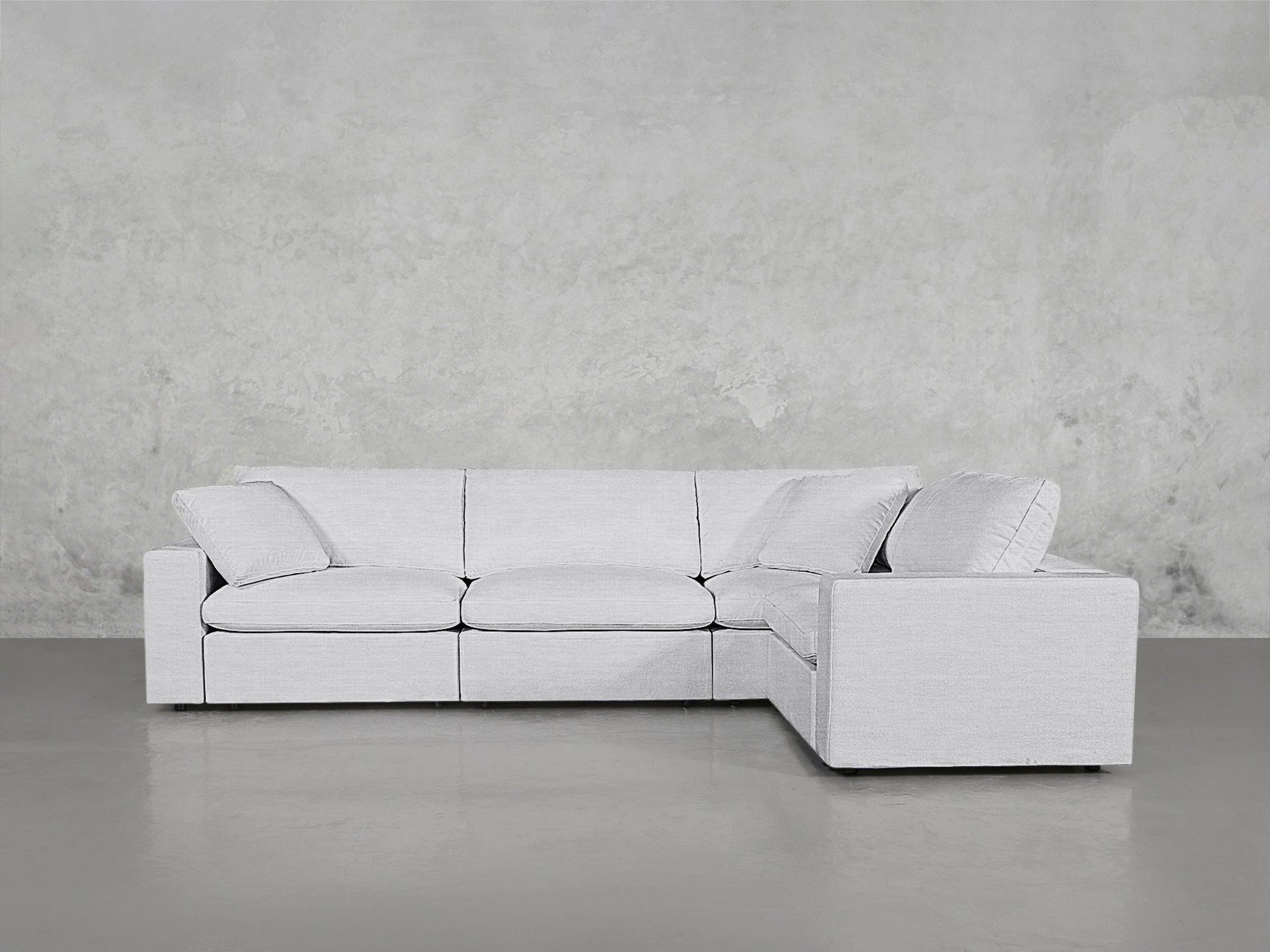 The 4-Seat Modular Corner Sectional, featuring three seat cushions, wide armrests, and back and side pillows for comfort, is shown in light gray on a smooth gray floor against a textured gray wall.