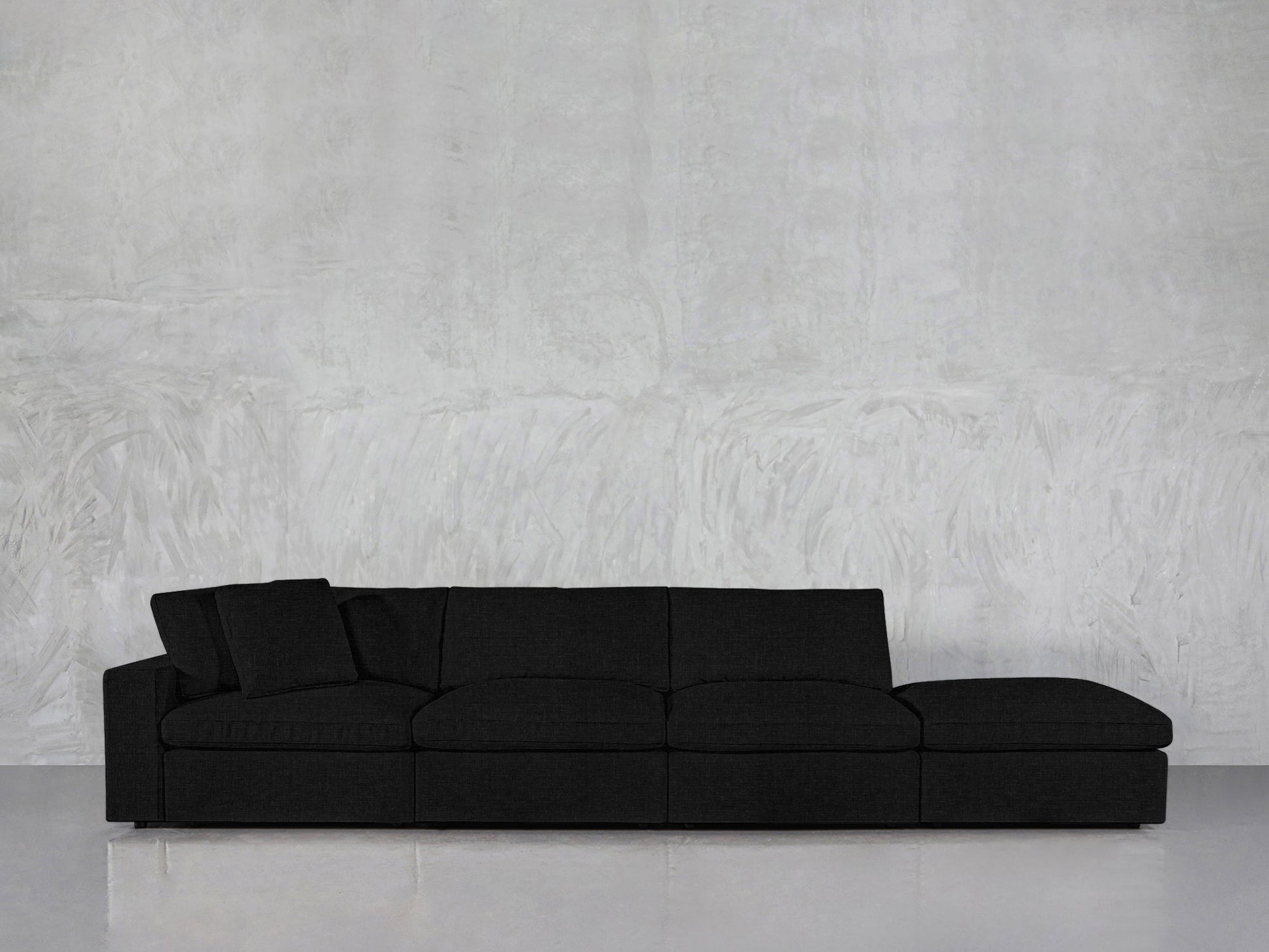 The 4-Seat Modular Lounger Sofa in dark gray features four seat sections and a matching throw pillow, displayed against a light gray textured wall and a light floor.