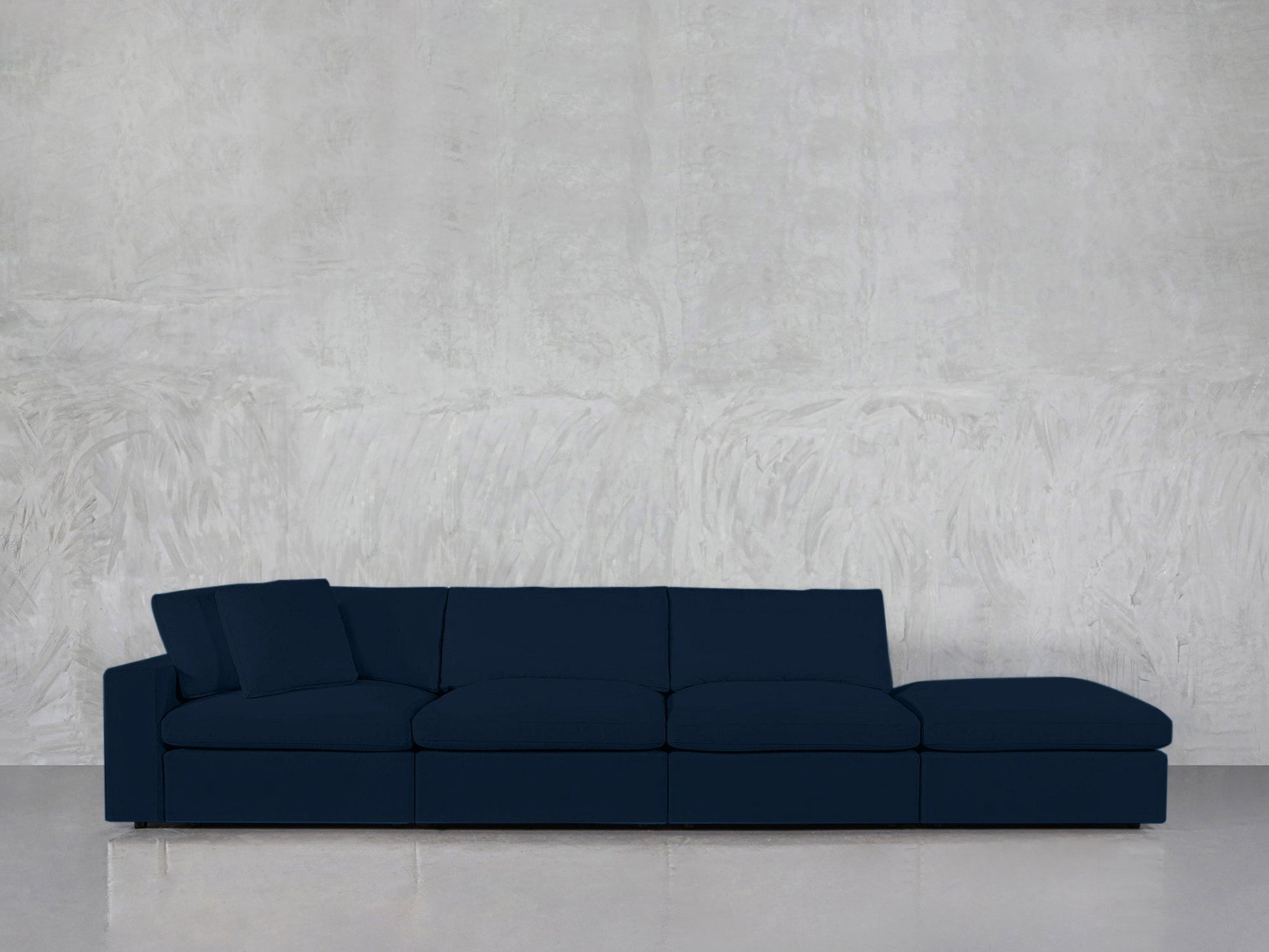 A modern, dark blue 4-Seat Modular Lounger Sofa with two back pillows is placed against a light gray textured wall on a reflective floor.