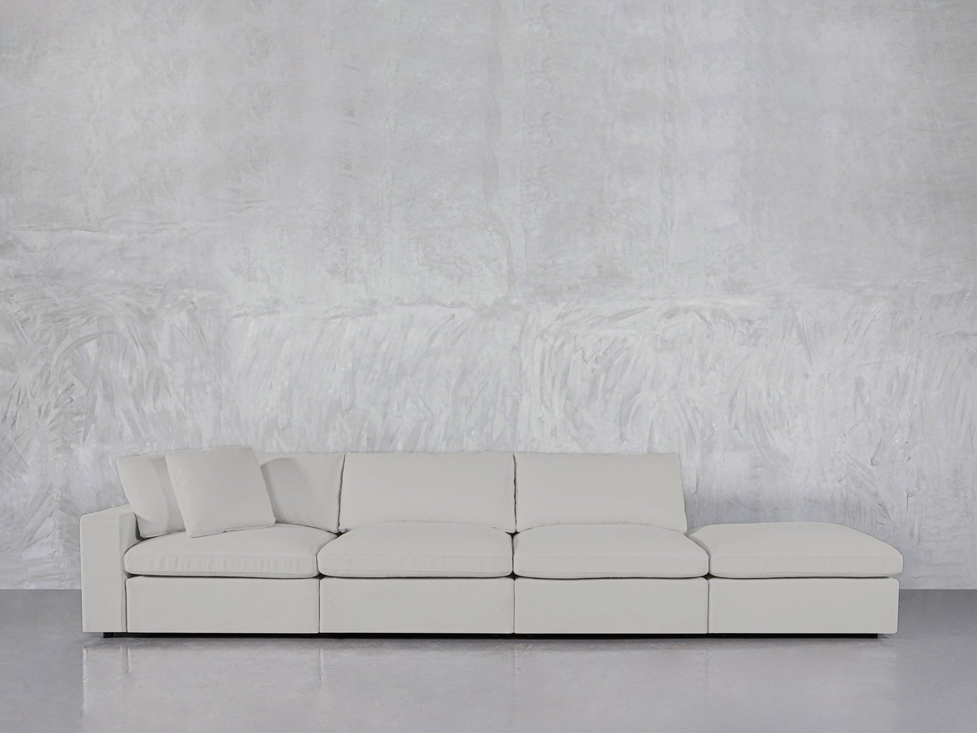 The 4-Seat Modular Lounger Sofa, in light gray with three seat cushions, a right-side chaise lounge, and matching pillows, sits against a textured gray wall on a shiny floor.