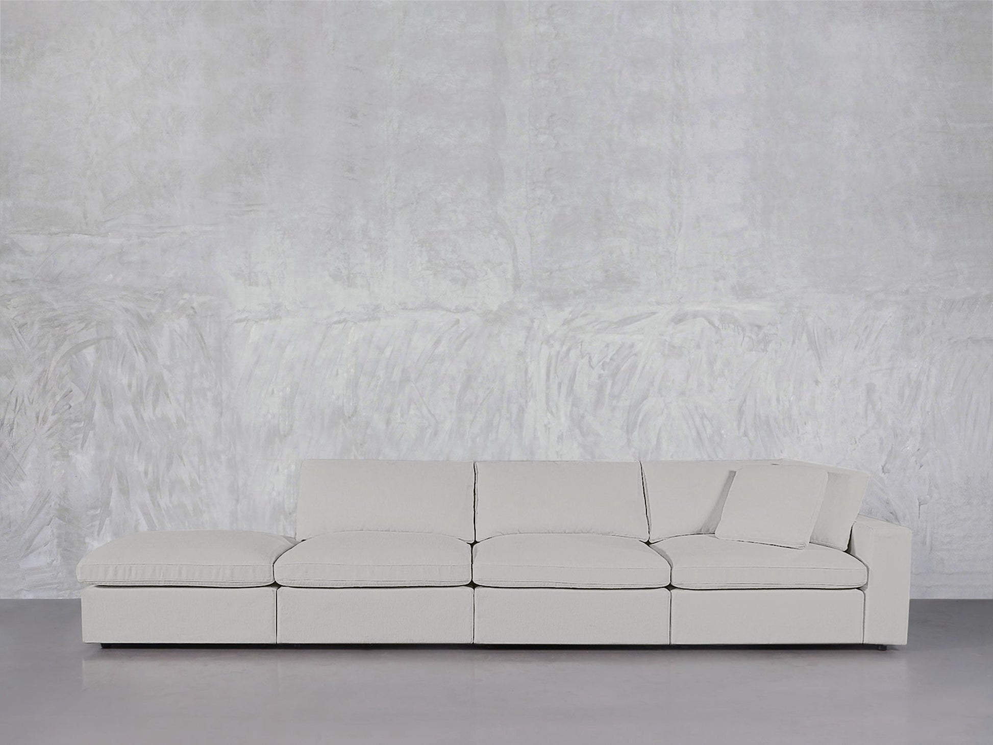 The 4-Seat Modular Lounger Sofa, in light gray with one right-side cushion, stands against a textured light gray wall and smooth gray flooring.