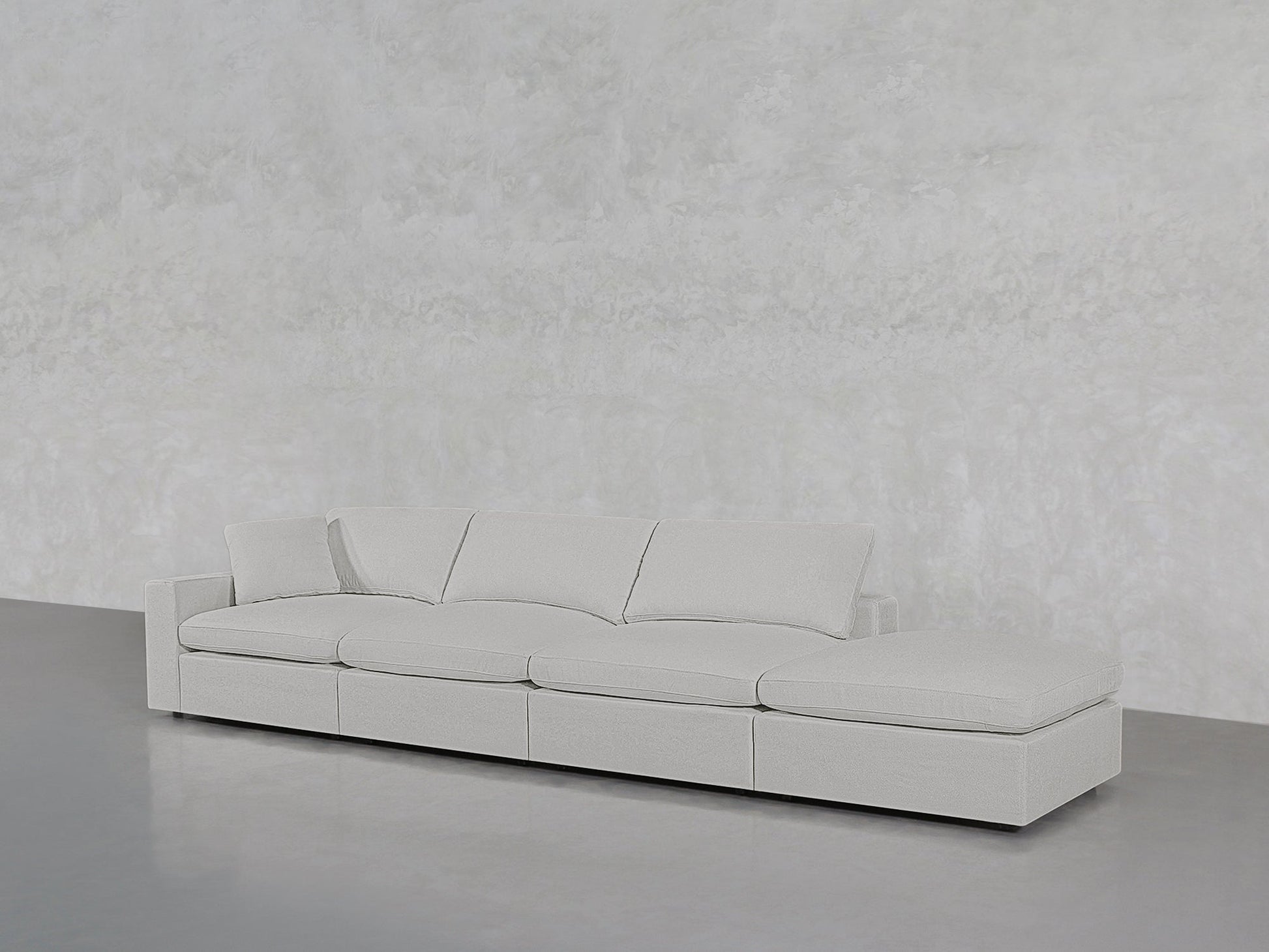 The 4-Seat Modular Lounger Sofa, in modern light gray with four cushions, is displayed against a plain textured light gray wall and a smooth gray floor.