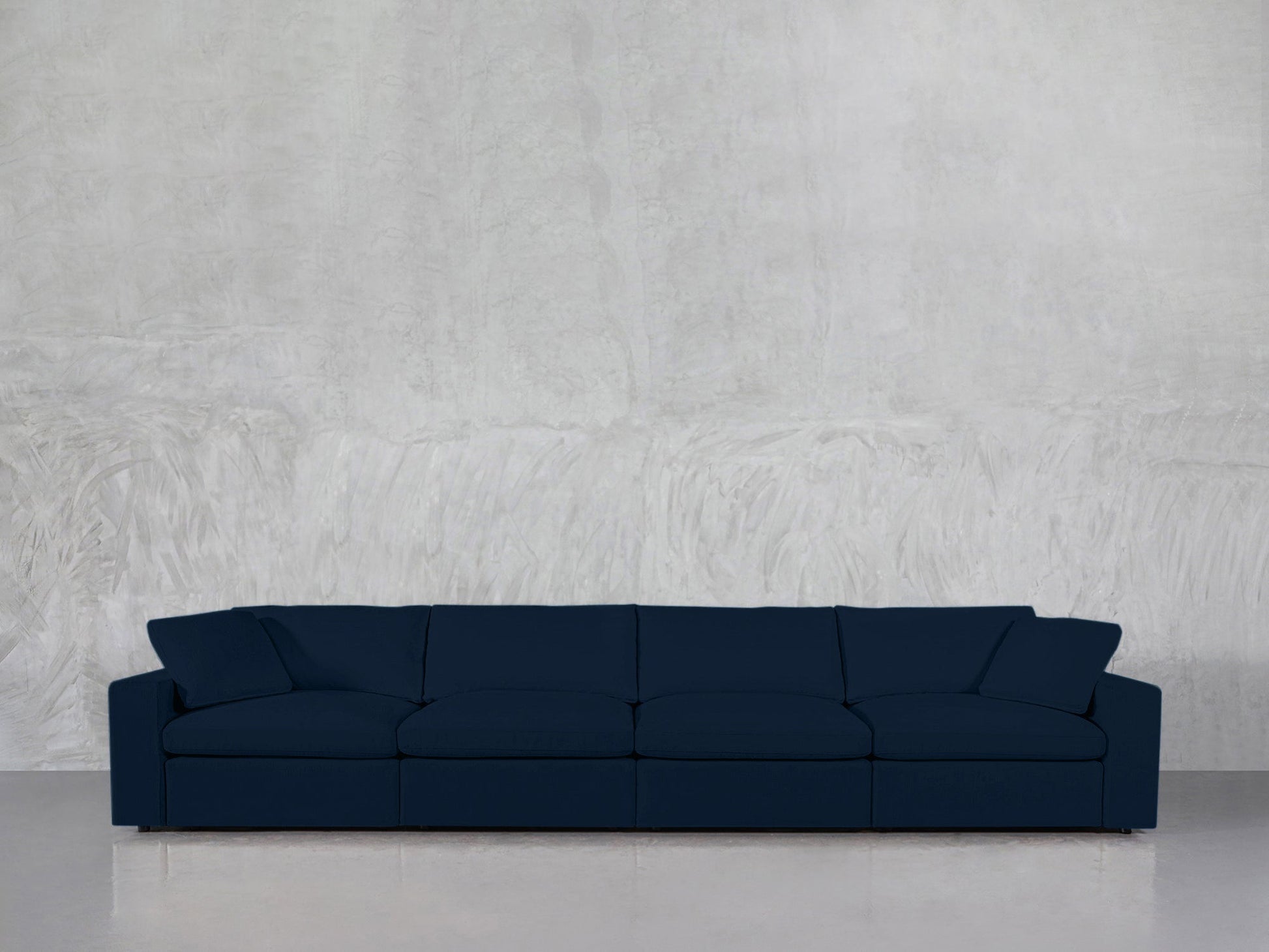 A 4-Seat Modular Sofa in dark blue with four seat cushions and two side pillows is placed against a light gray textured wall on a shiny floor.