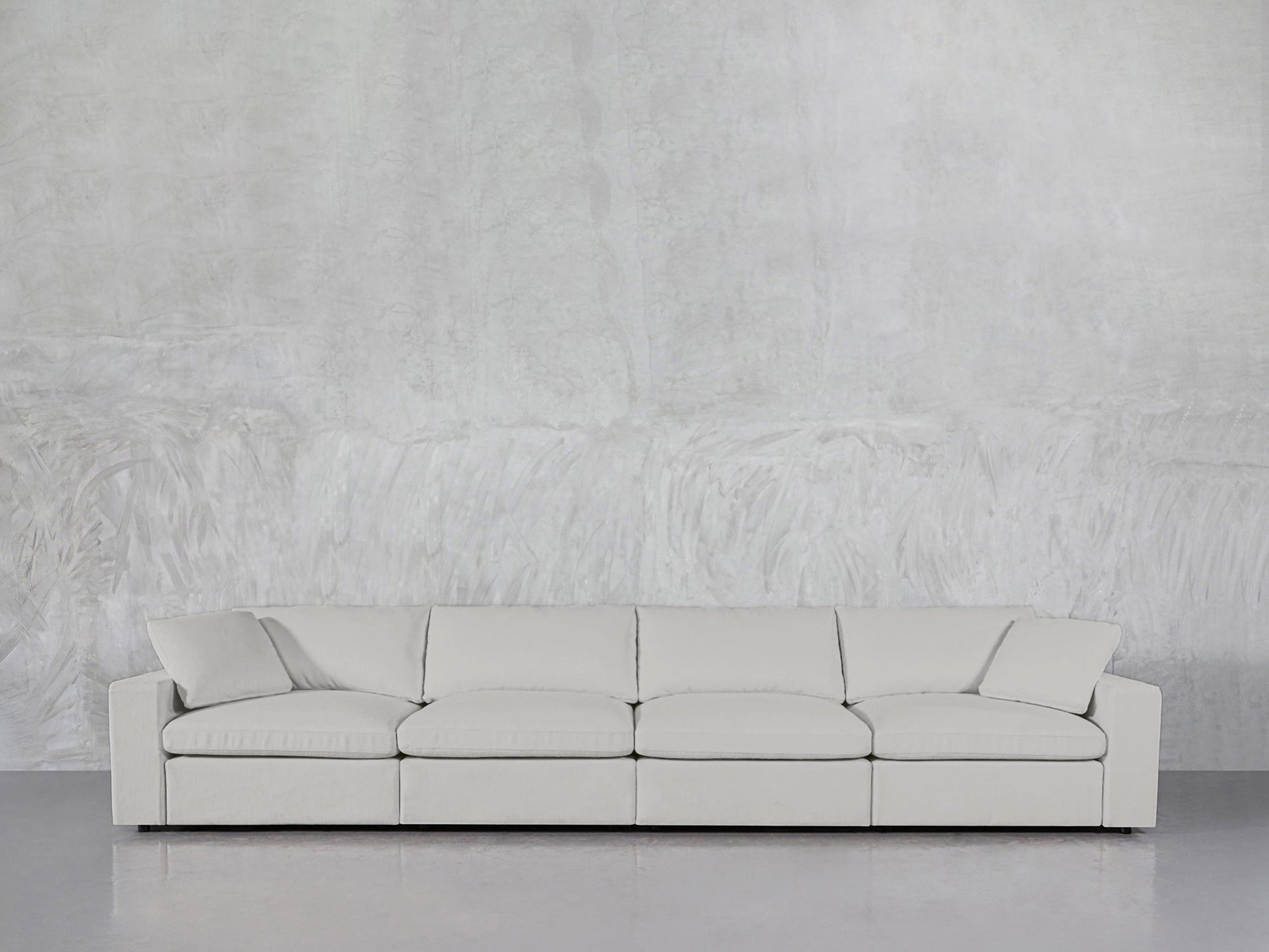 A modern 4-Seat Modular Sofa with square arms and five back cushions in light gray rests against a textured, light wall on a smooth, reflective floor.