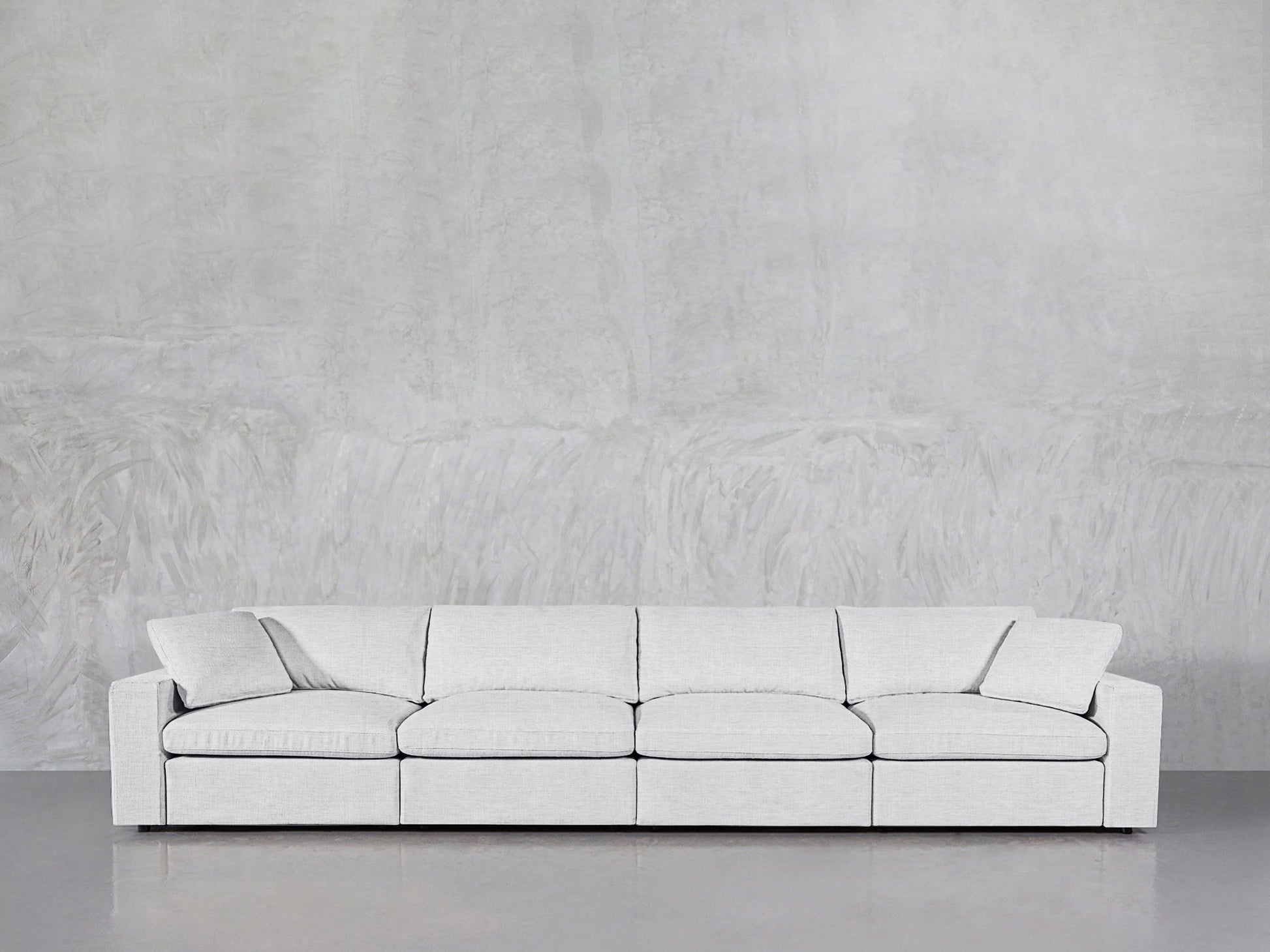 A modern 4-Seat Modular Sofa in light gray with cushions and armrests is placed against a textured gray wall and a smooth gray floor.