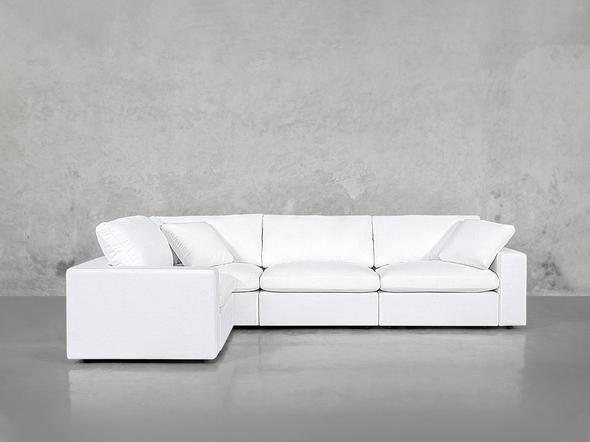 A 4-Seat Modular Corner Sectional with clean lines and a modern white finish is set against a plain, light gray concrete wall and floor.