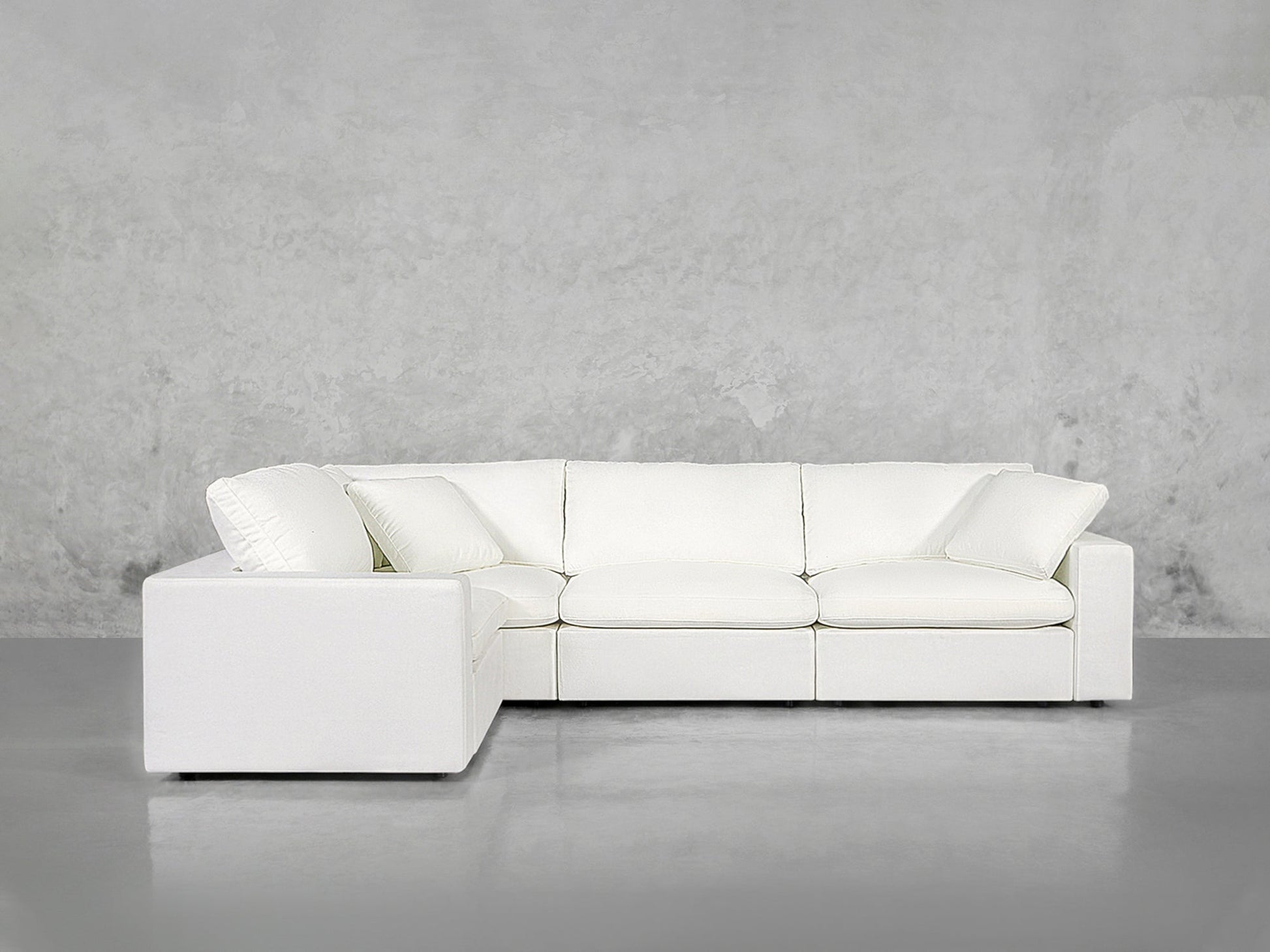 A modern 4-Seat Modular Corner Sectional in white, featuring three seat and back cushions, set against a plain light gray concrete wall and floor.
