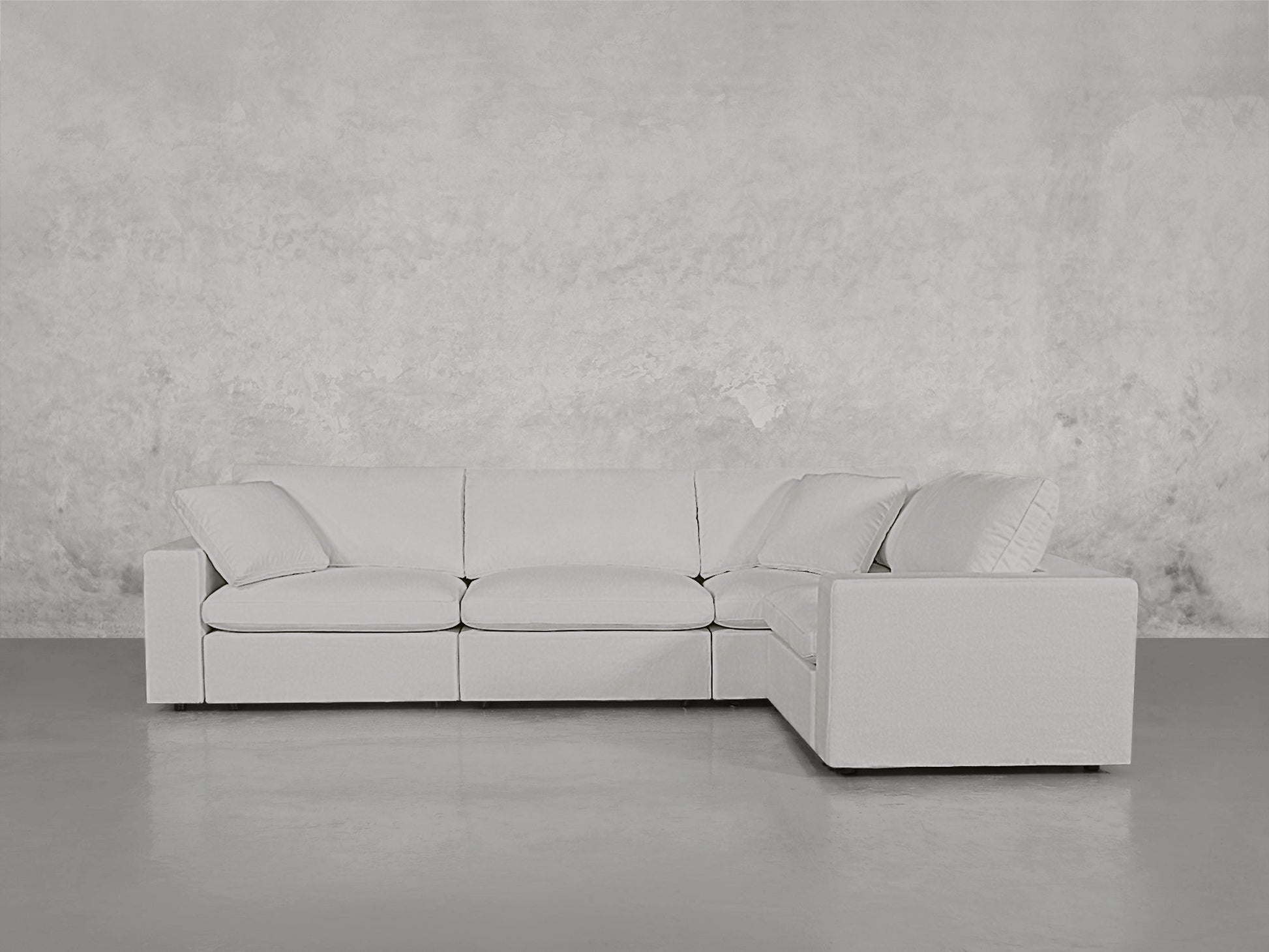 A 4-Seat Modular Corner Sectional in light gray with clean lines sits on a polished concrete floor against a textured, pale gray wall, featuring three seat cushions and an L-shaped design.