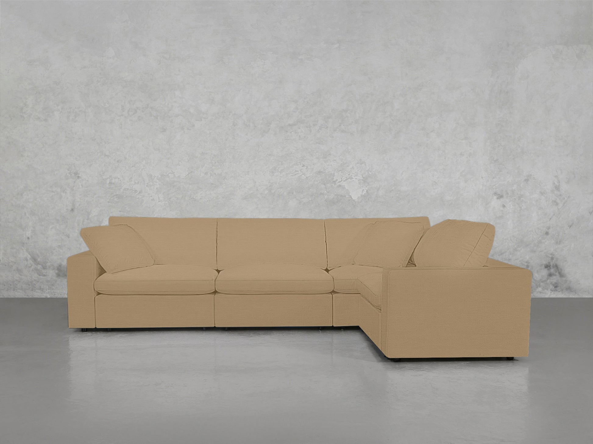 A 4-Seat Modular Corner Sectional in beige with cushions and wide armrests is set against a textured light gray concrete wall on a smooth gray floor.