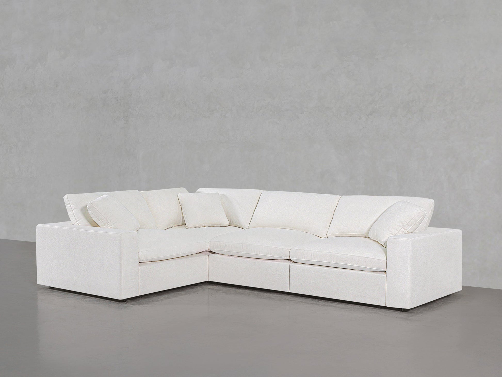 A 4-Seat Modular Corner Sectional with plush cushions and armrests, featuring an L-shaped white design, is set against a plain light gray wall on a smooth gray floor.