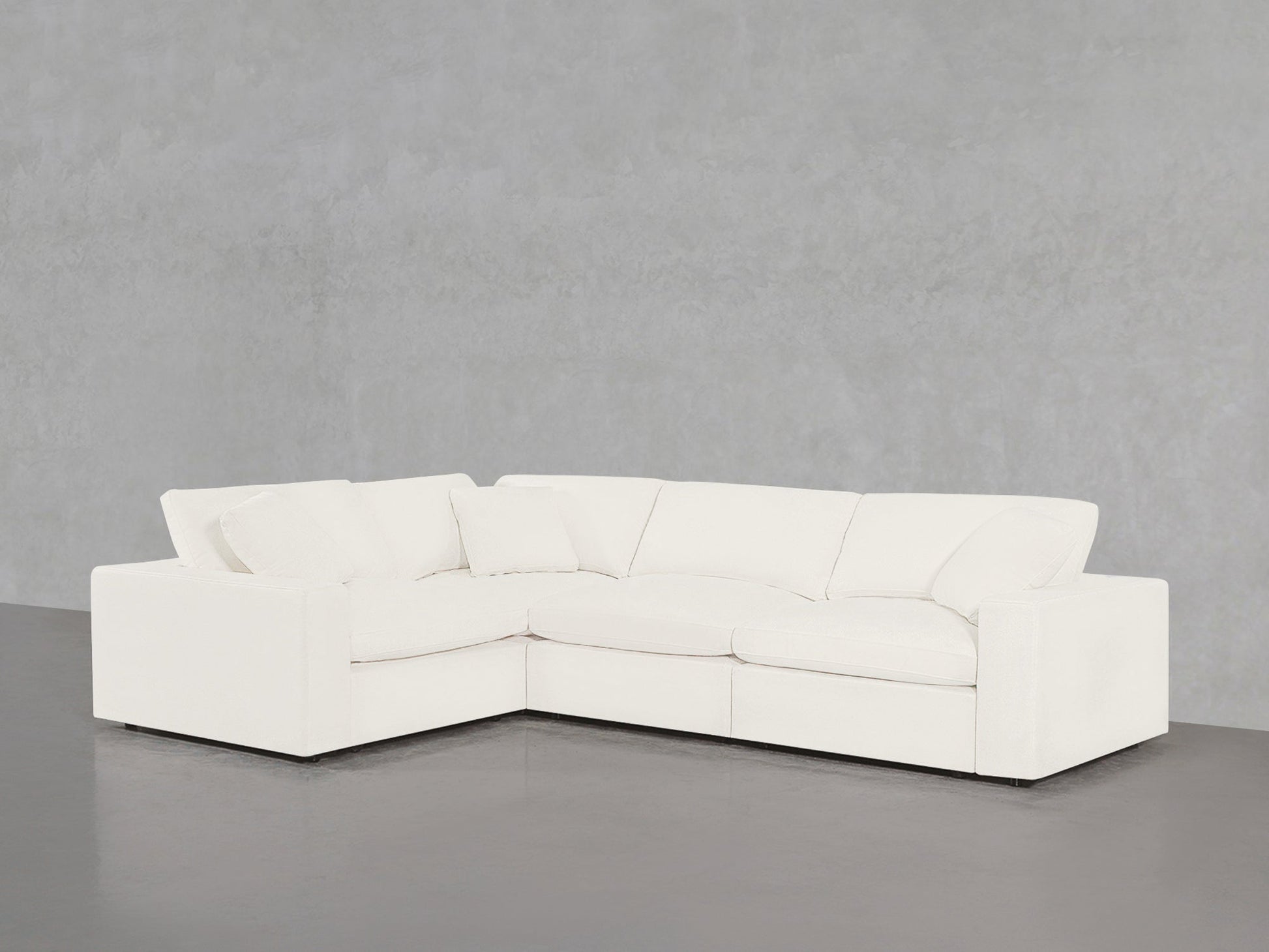 A 4-Seat Modular Corner Sectional in white with three seat and three back cushions, set in a minimalist room featuring a plain gray wall and a dark gray floor.