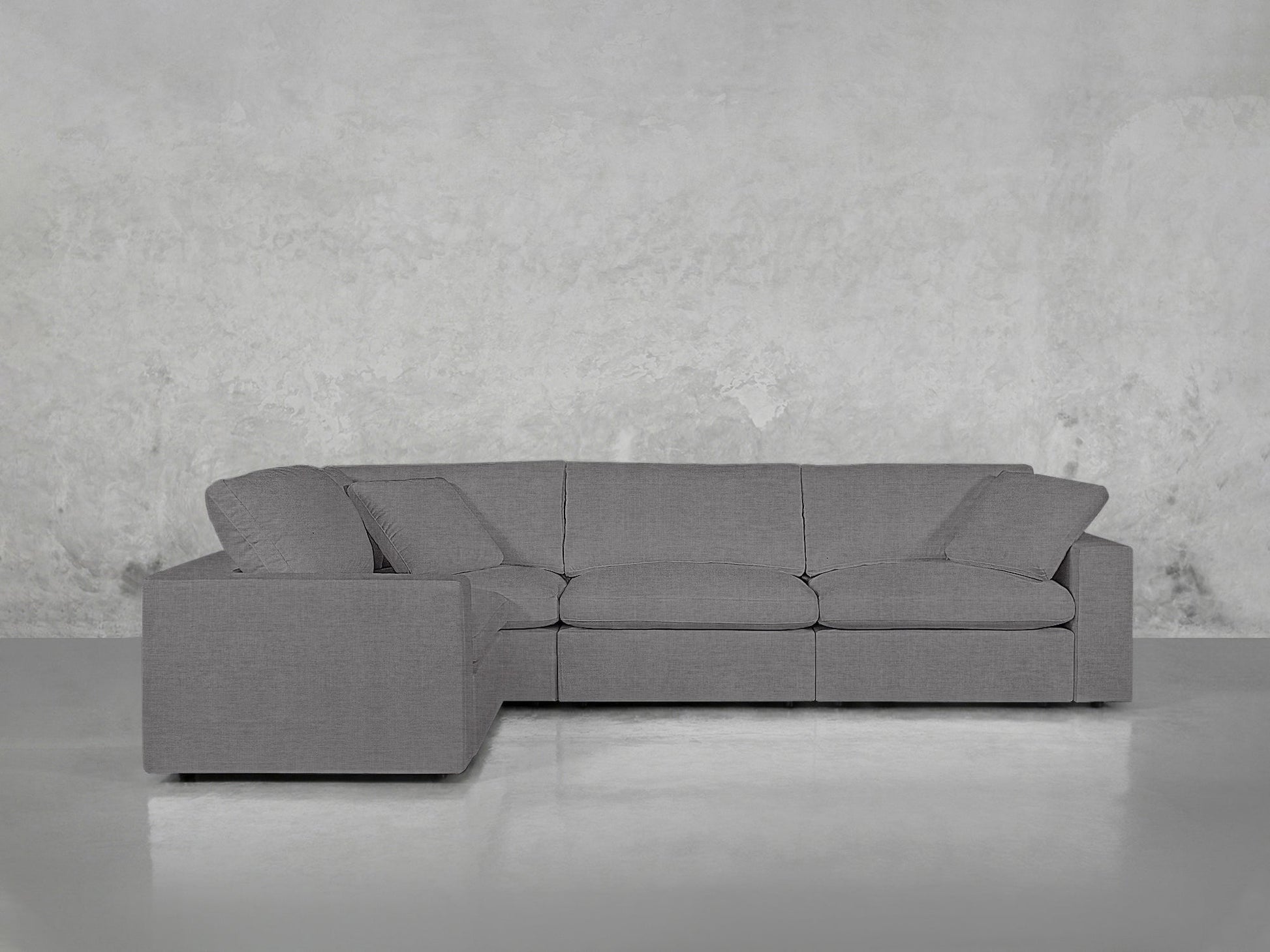 A 4-Seat Modular Corner Sectional in modern gray is set against a textured light gray wall on a glossy floor, with matching three seat and back cushions.