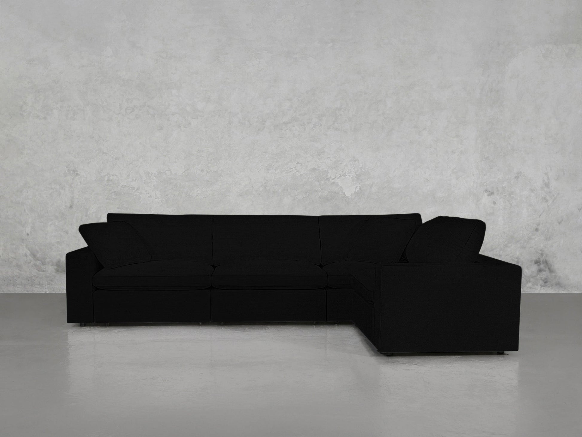 A 4-Seat Modular Corner Sectional with modern black upholstery and clean lines is set in a minimalist room featuring a polished concrete floor and a light gray textured wall.